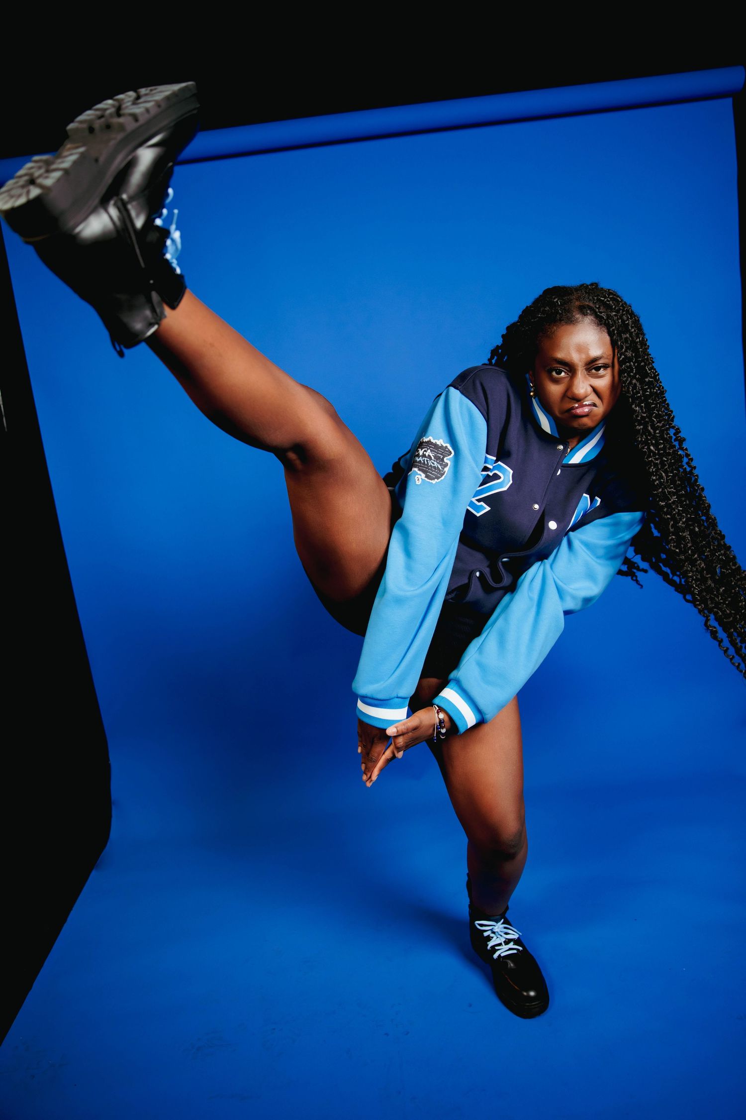 Athletic woman in blue varsity jacket performing high kick pose on blue studio background wearing black shoes.
