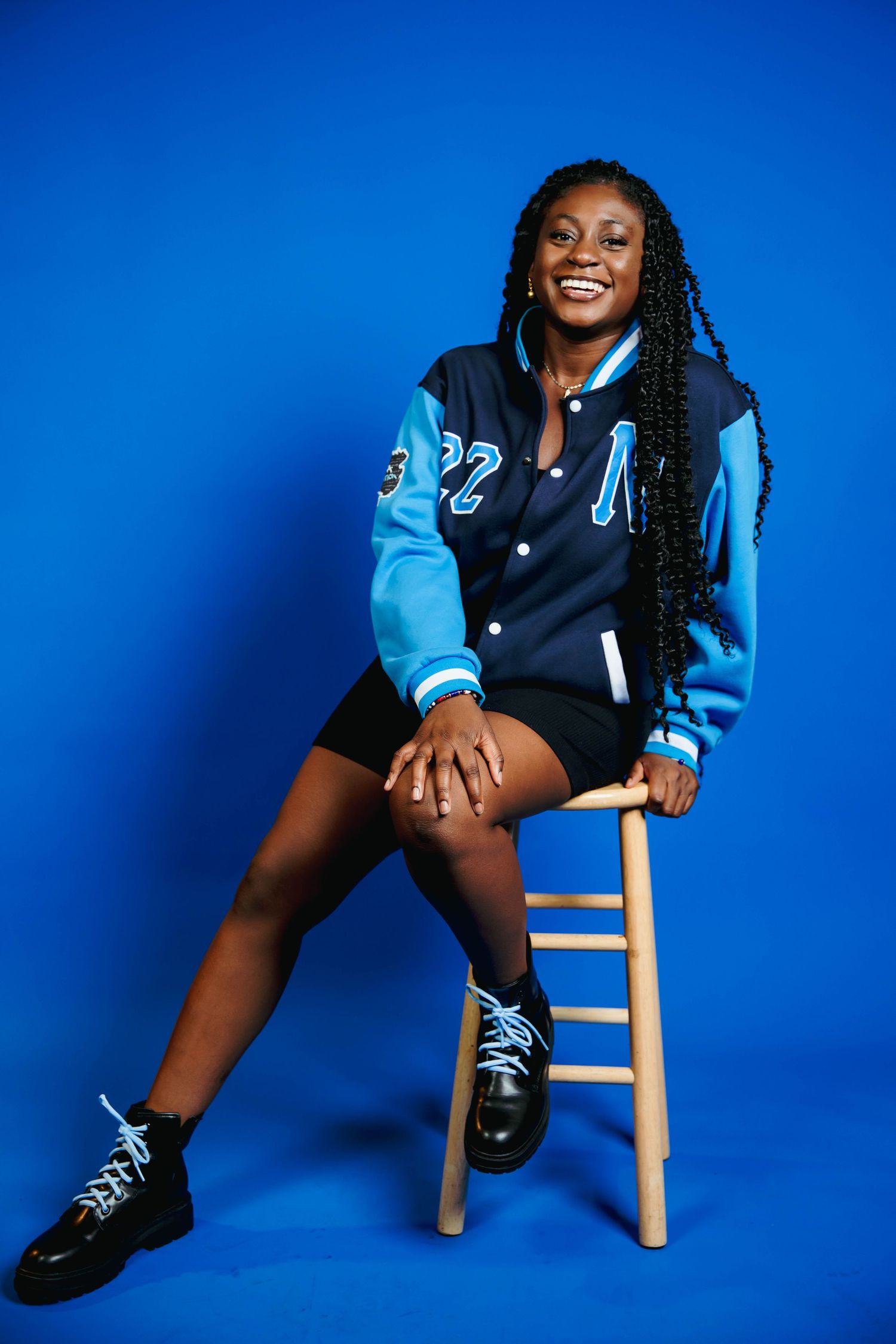 Smiling young woman in blue varsity jacket sitting on wooden stool against bright blue background.