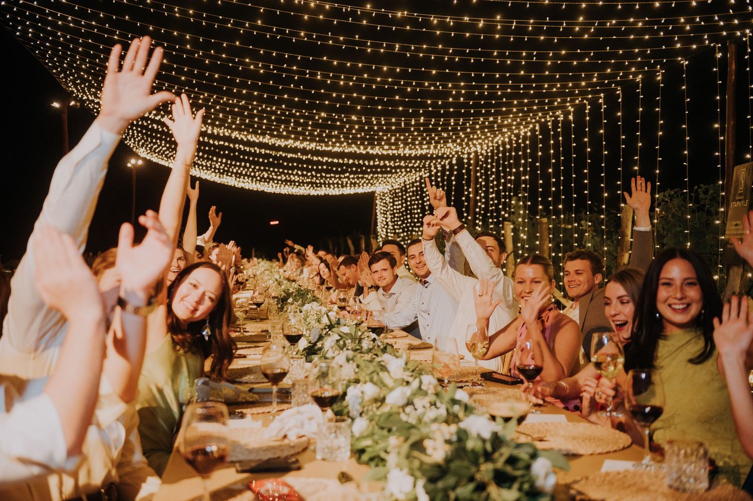Joyful wedding guests raise hands at long dinner table adorned with greenery under string lights at night.