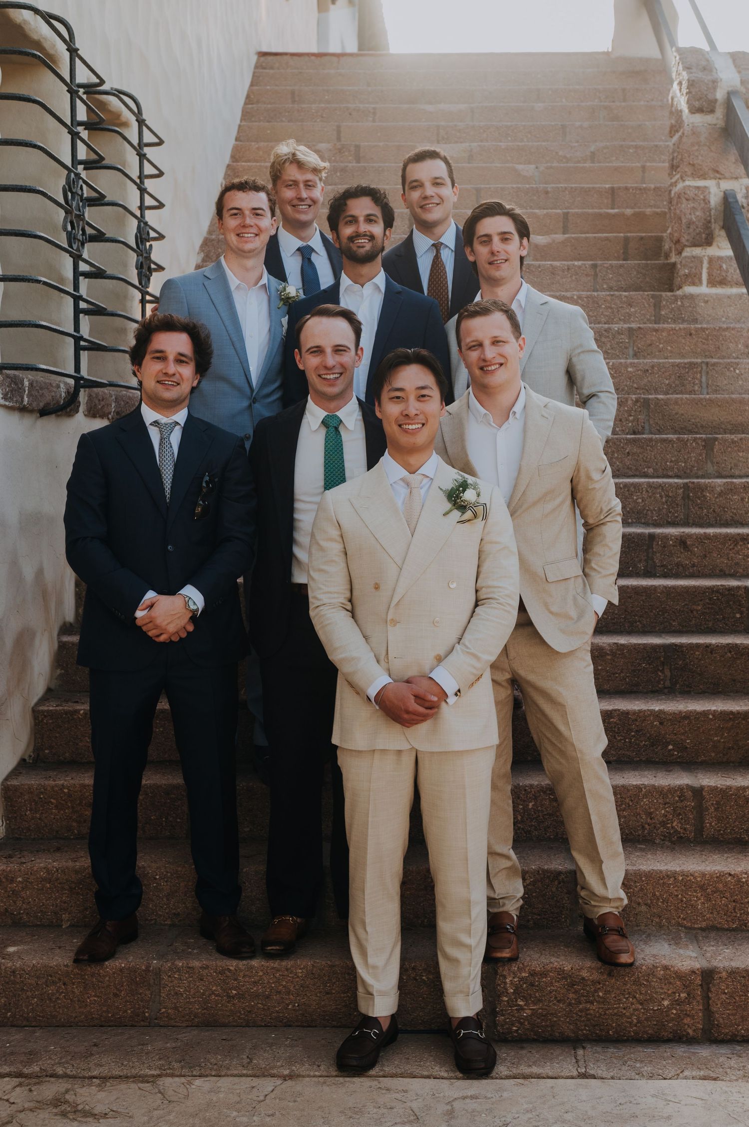 Groom in cream double-breasted suit posing with groomsmen on stone staircase.