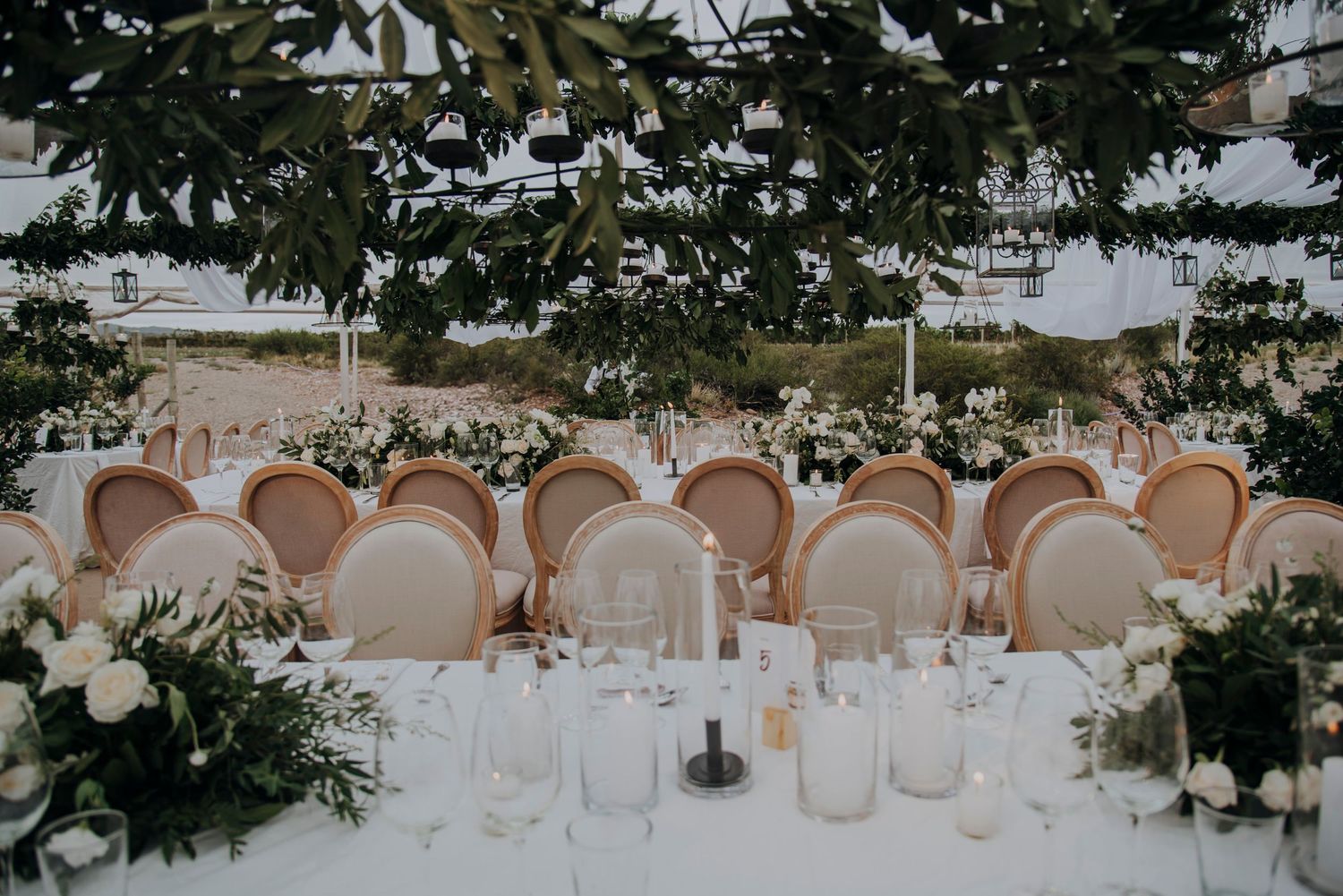 Wedding reception tables with white linens, candles, floral centerpieces under leafy canopy.