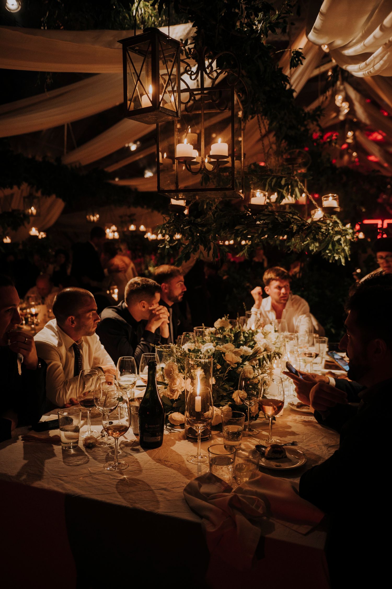 Guests dining by candlelight at romantic wedding reception with hanging lanterns and greenery.