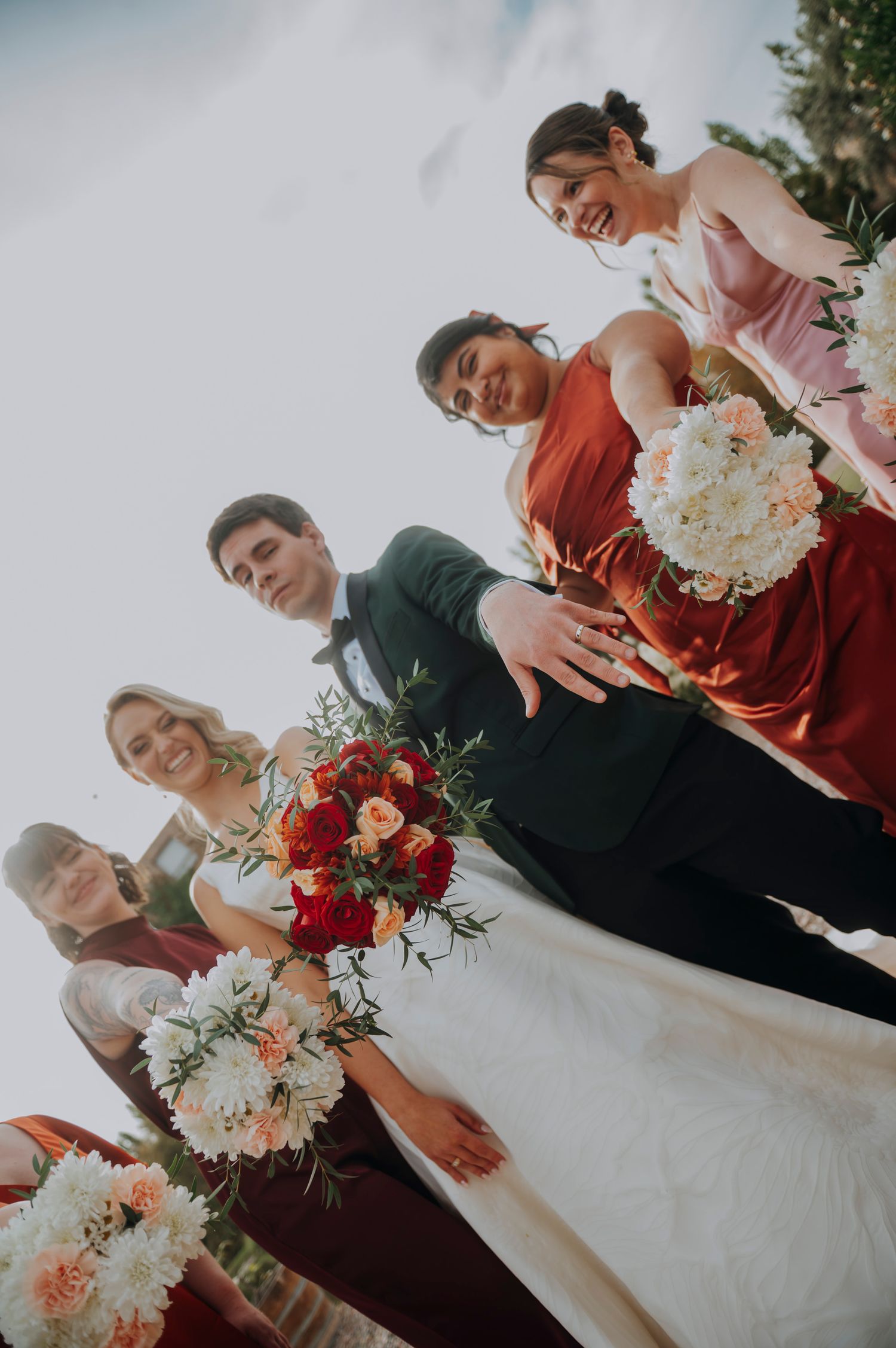 Low-angle wedding party photo featuring bride in white gown, groom in green suit, and bridesmaids in red holding bouquets.