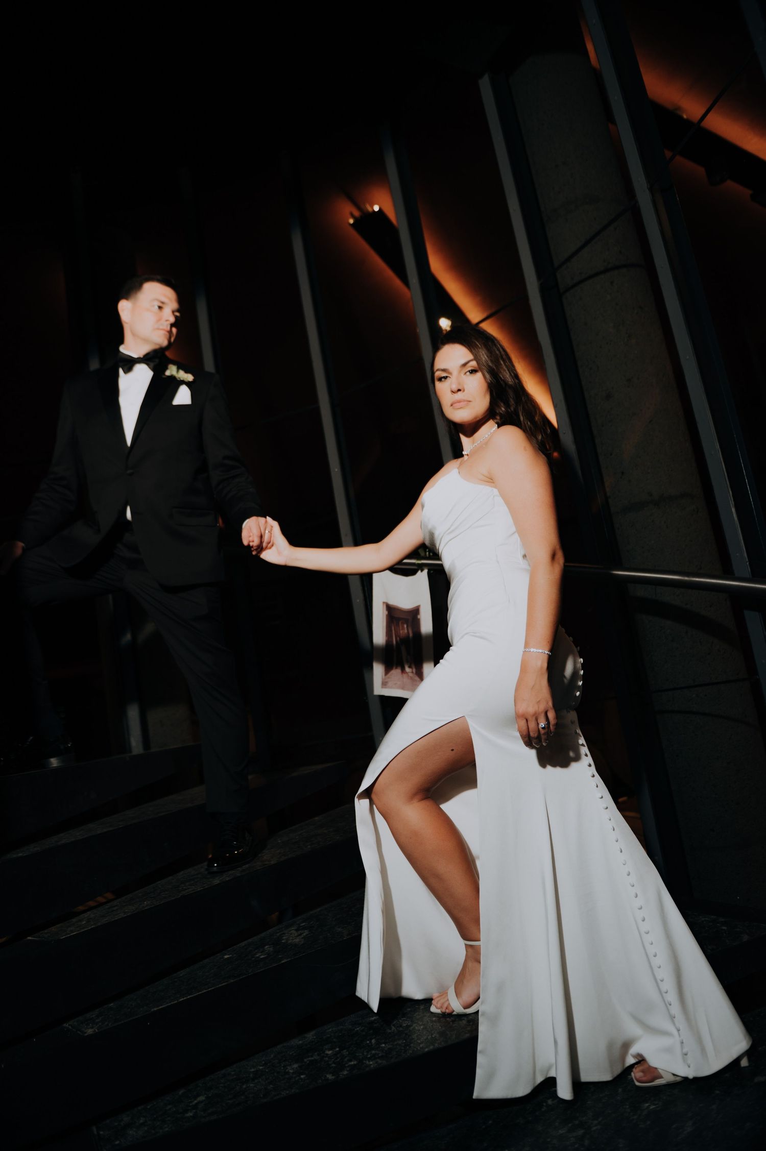Elegant bride in sleek white gown holds grooms hand on dark staircase at night during wedding reception.
