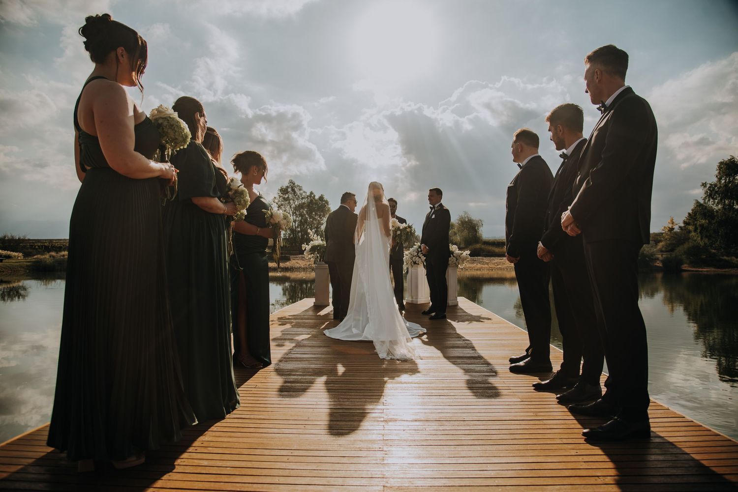 Wedding party standing on sunlit dock over lake during outdoor ceremony with bridesmaids in sage green dresses.