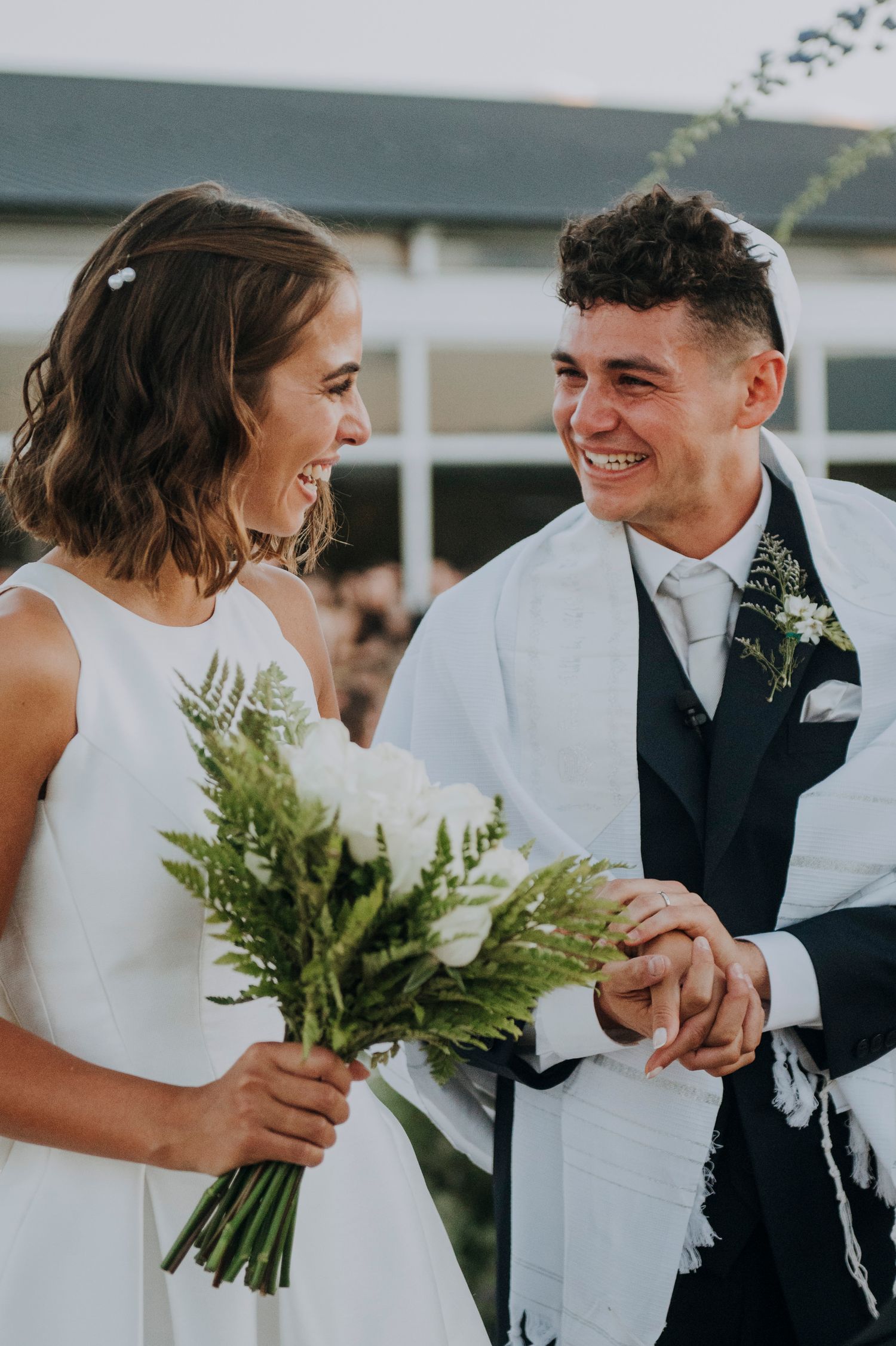 Smiling bride and groom exchange vows outdoors, bride holding white rose and fern bouquet.