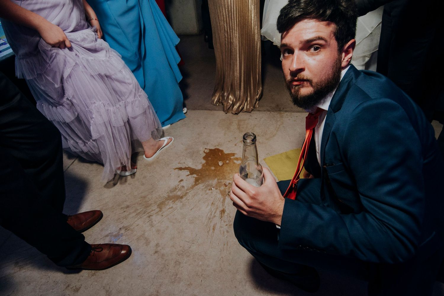 Wedding guest in blue suit sitting on floor holding bottle near spilled drink.