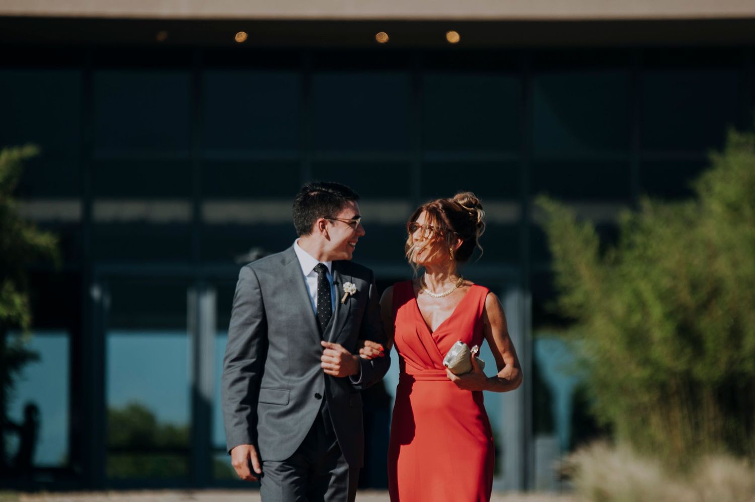 Elegant couple in formal attire smiling at each other outdoors, woman in red dress, man in gray suit with boutonniere.