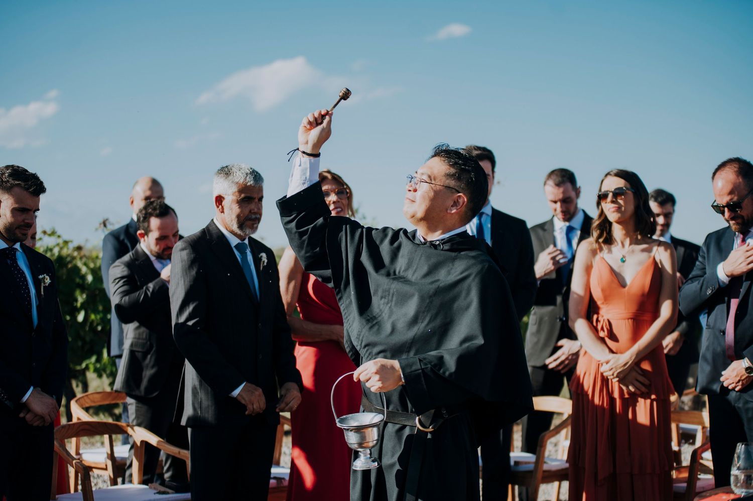 Officiant raises a ceremonial aspergillum during an outdoor wedding ceremony with guests seated in rows.
