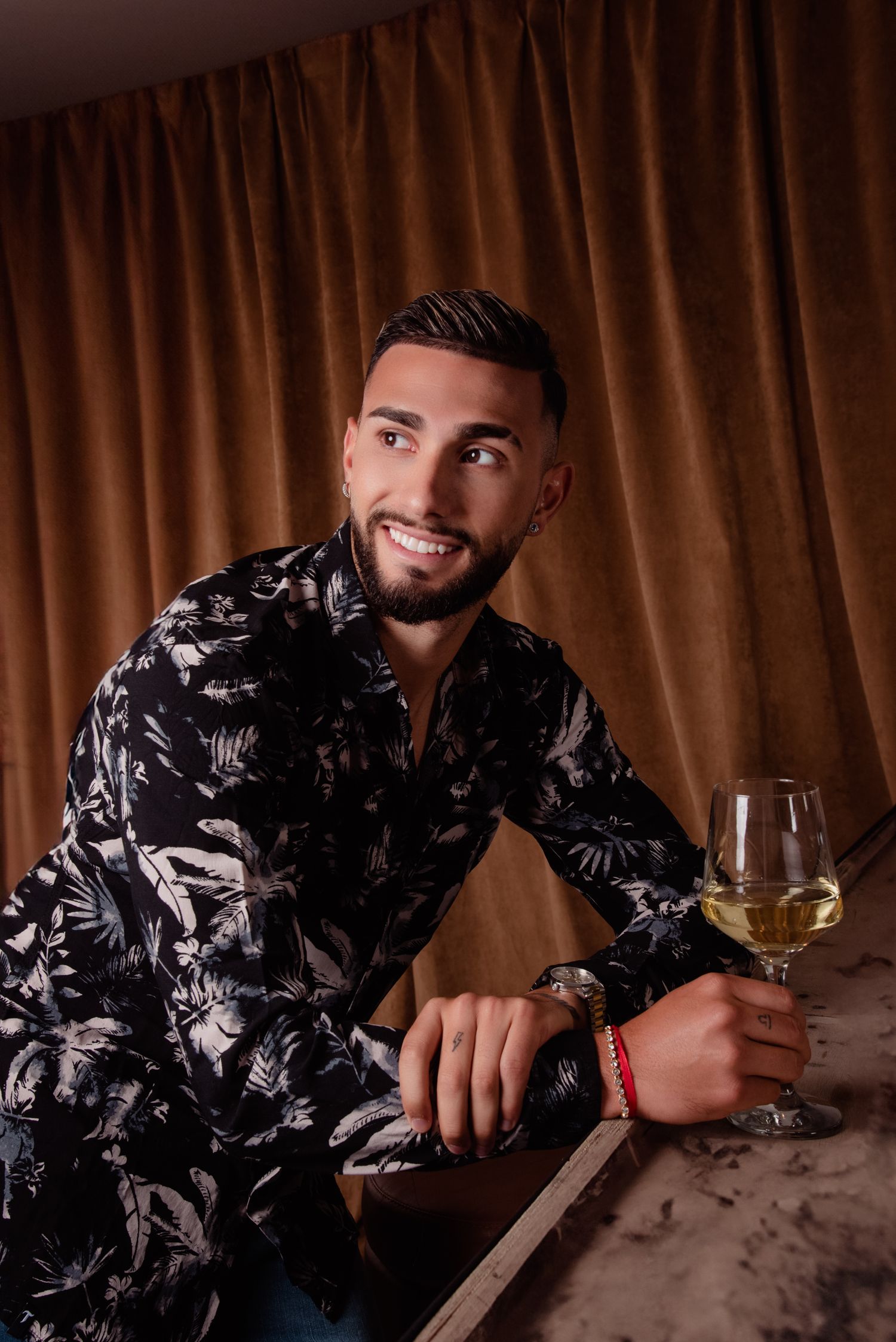 Taty Castellanos Smiling bearded man in floral shirt holding a wine glass at a table with velvet curtain backdrop.