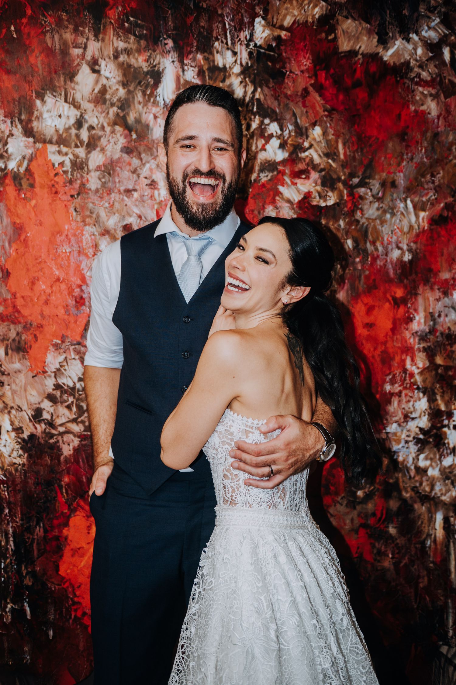 Laughing bride and groom embracing in front of vibrant red abstract painting backdrop. in anaia mendoza