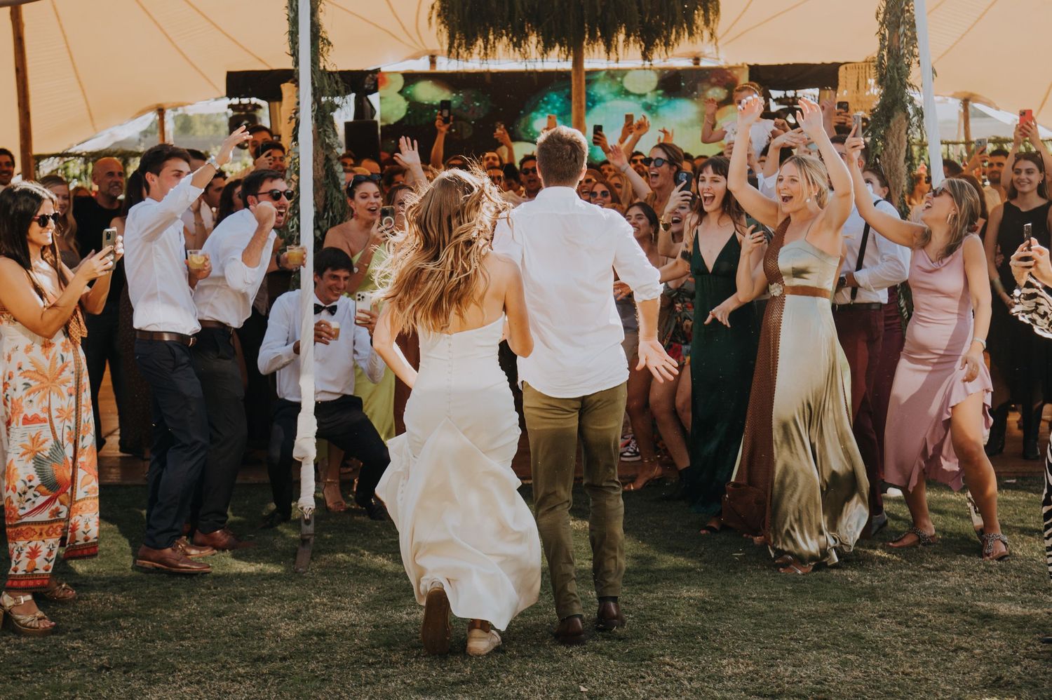 Bride and groom dance joyfully surrounded by cheering wedding guests under a tent at an outdoor reception.