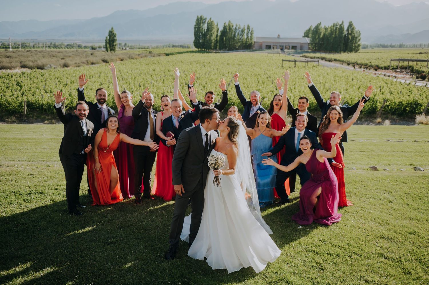 Wedding couple kissing while bridal party celebrates behind them at a vineyard with mountains in the background.
