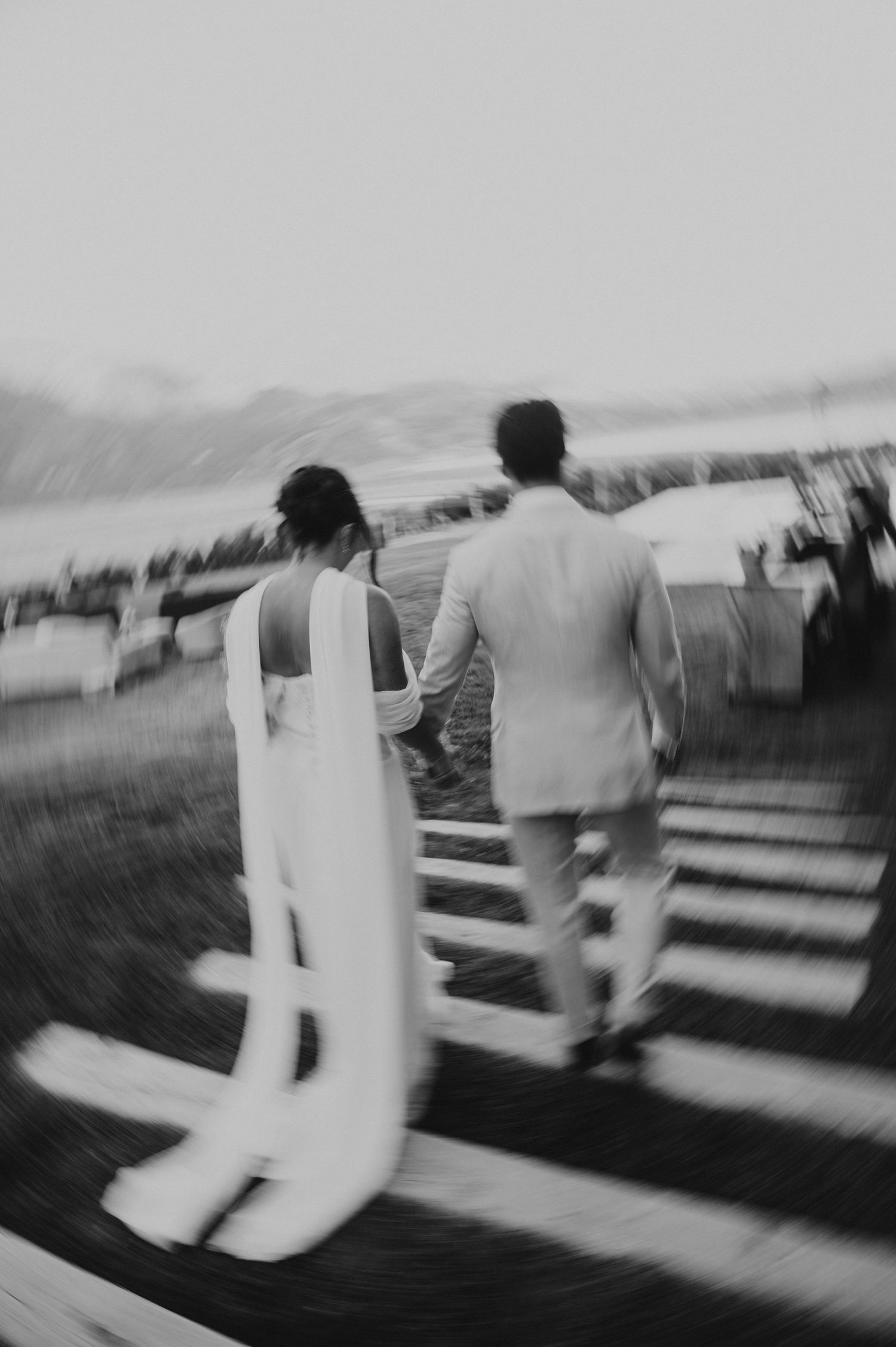 Black and white photo of a bride and groom holding hands, walking up stairs at an outdoor wedding venue.