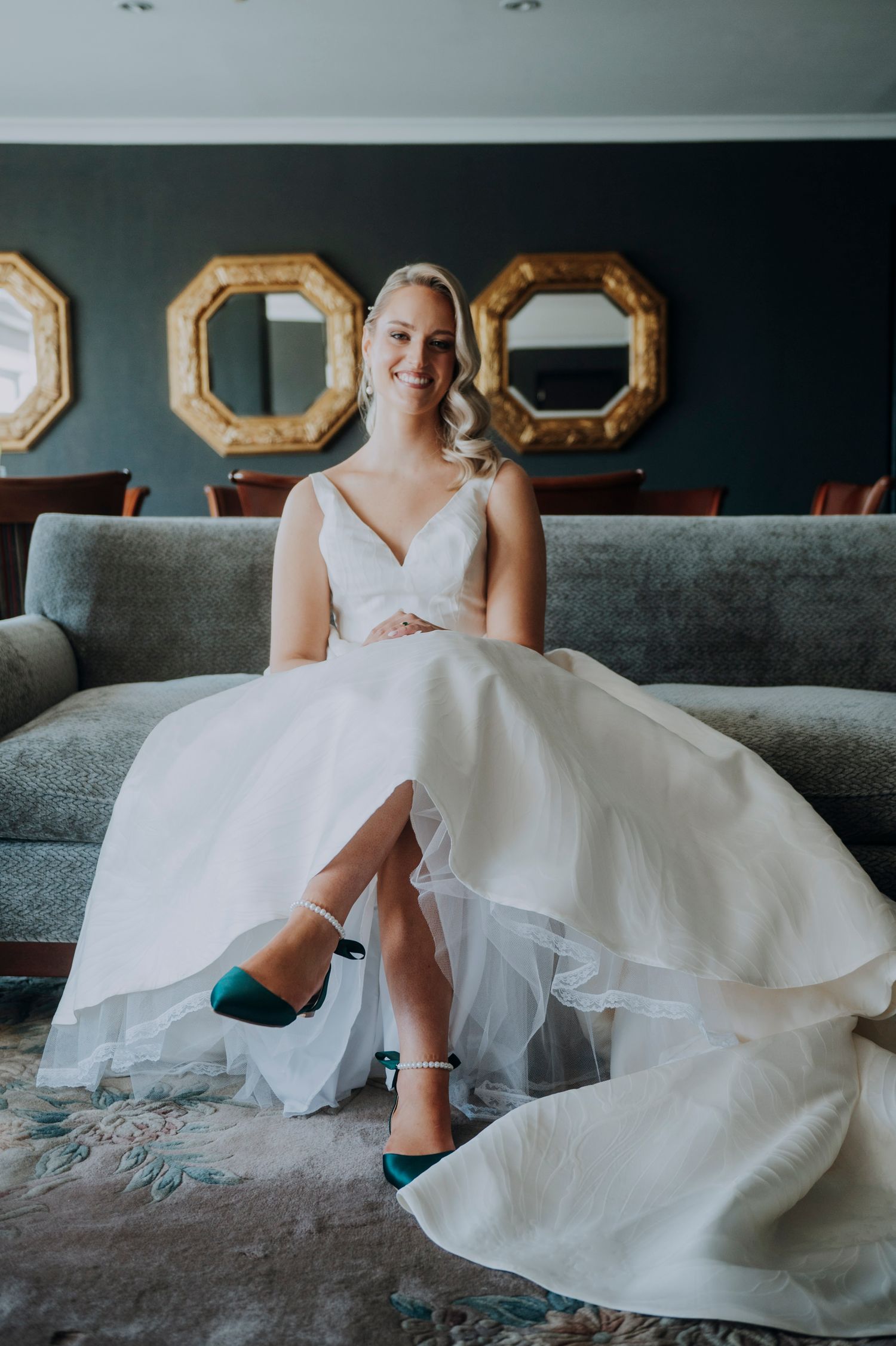 Bride in white ballgown with teal heels sits smiling on gray sofa with gold octagonal mirrors on wall.