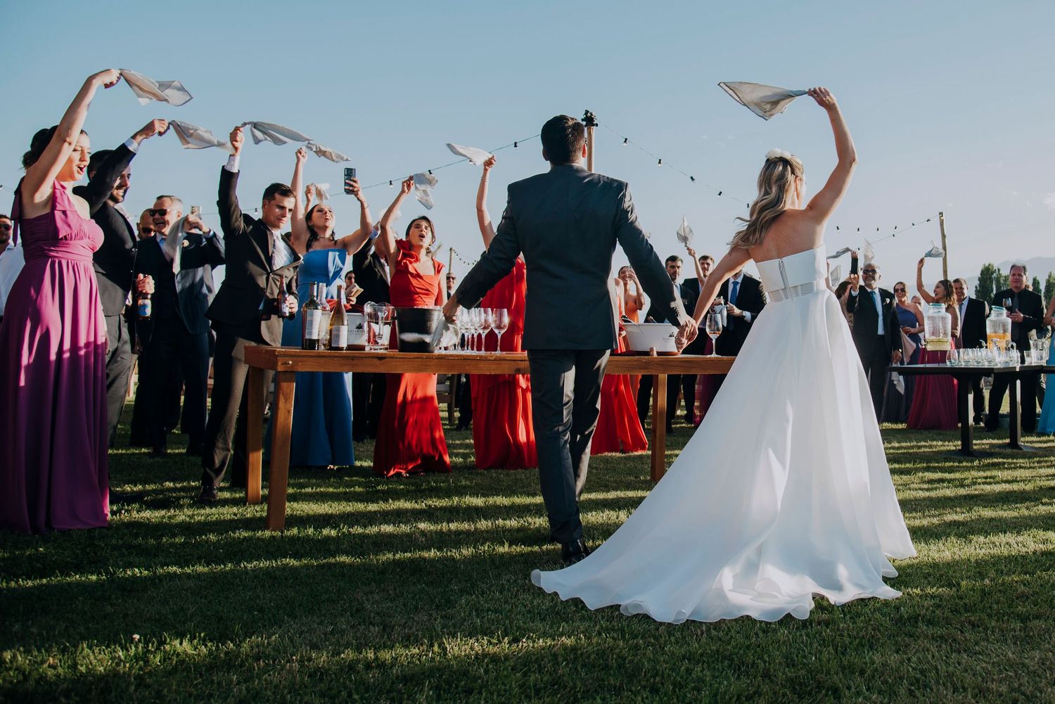 Bride and groom walk toward cheering guests waving white napkins at outdoor vineyard reception.