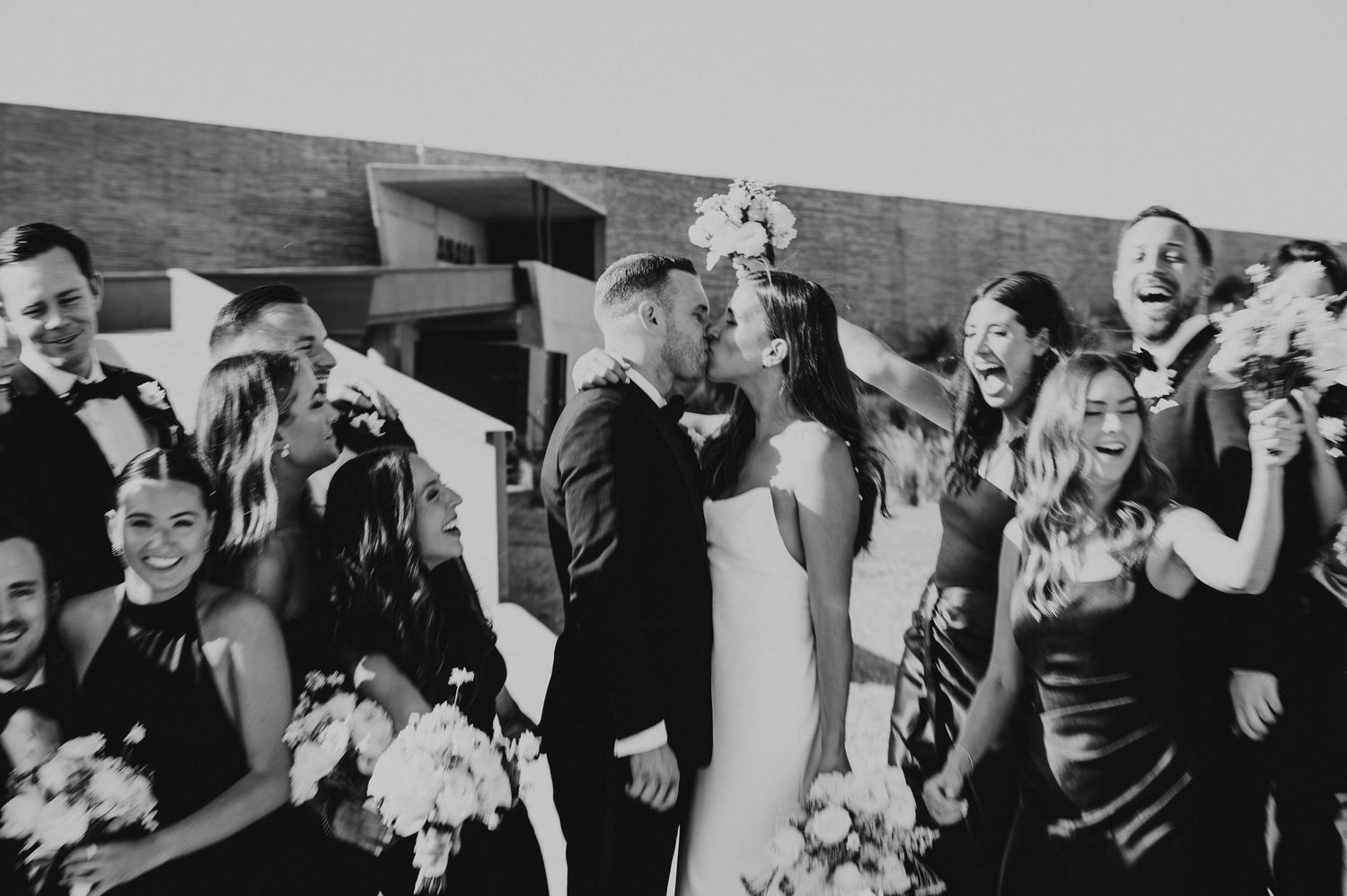 Wedding party celebrating with kiss in black and white photo surrounded by cheering guests.