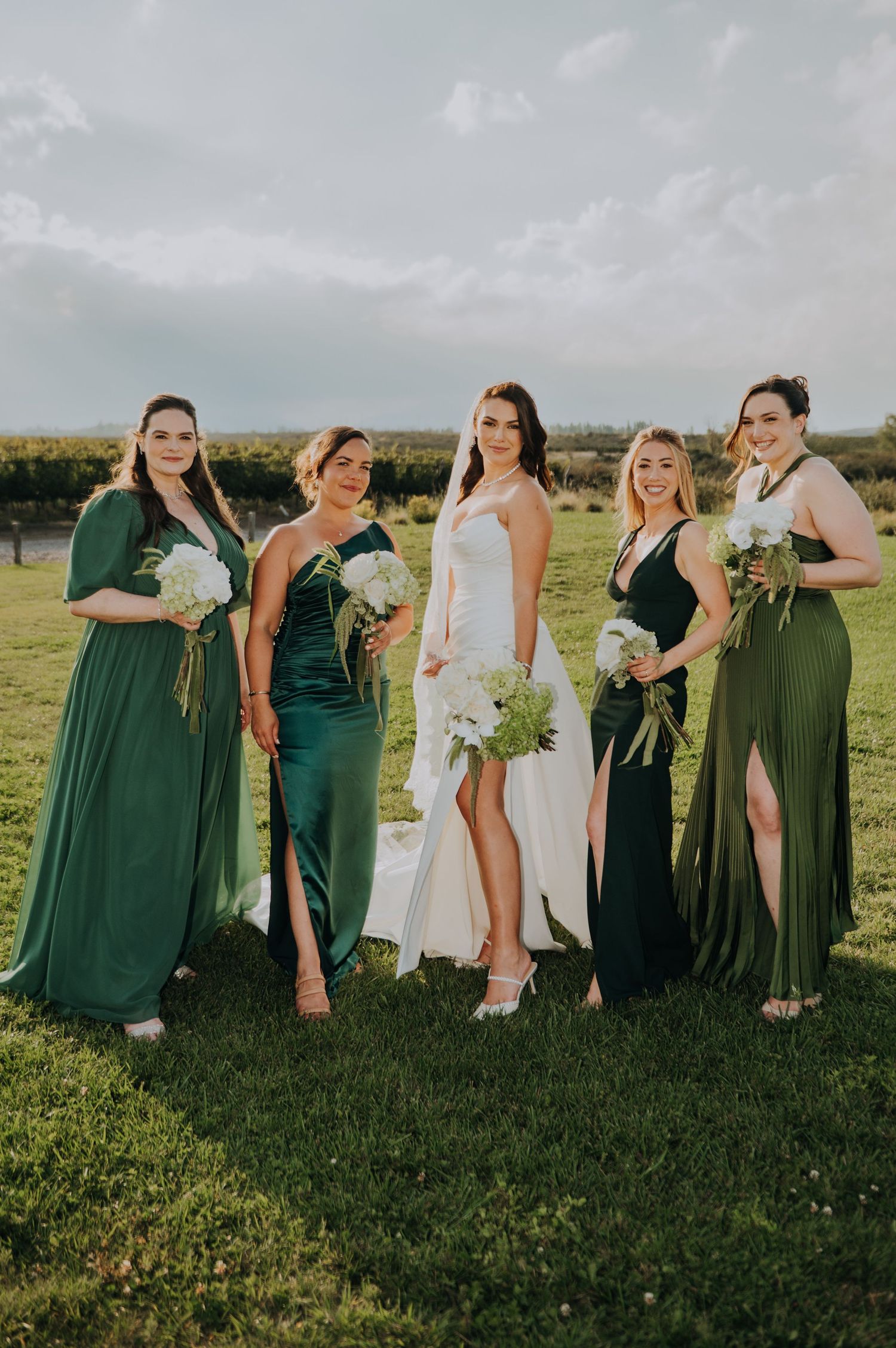 Bride in white gown poses with four bridesmaids in green dresses holding white bouquets on lush lawn.