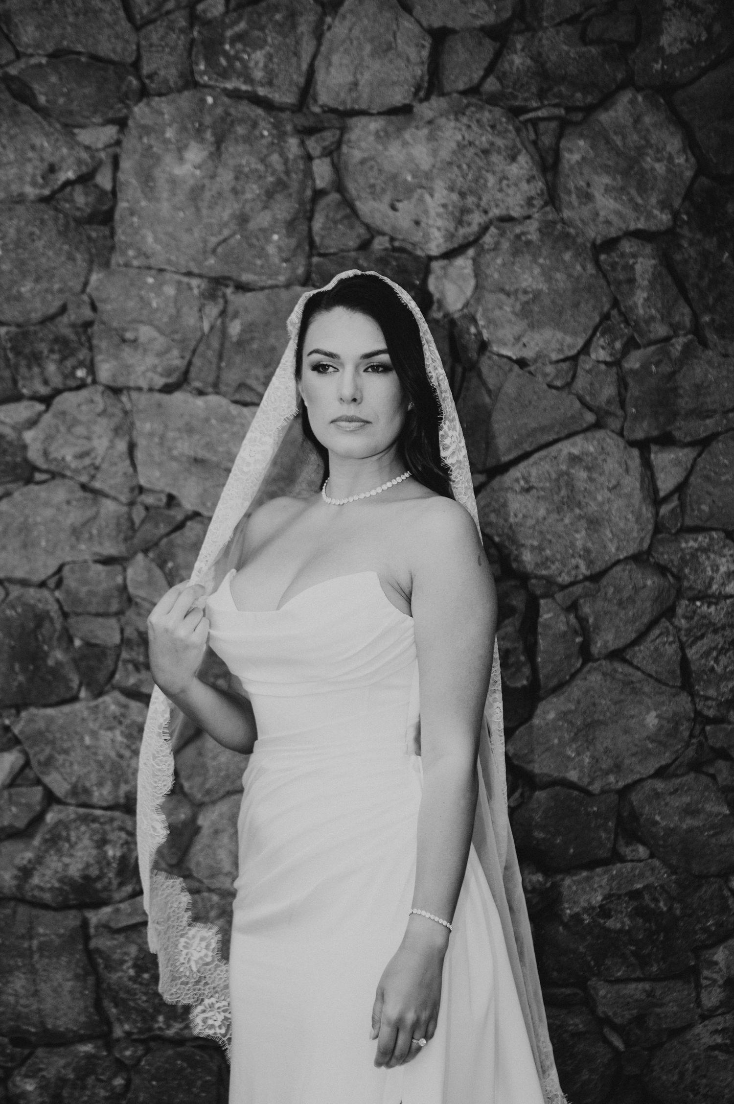 Elegant bride in strapless gown and lace veil poses against rustic stone wall in black and white.