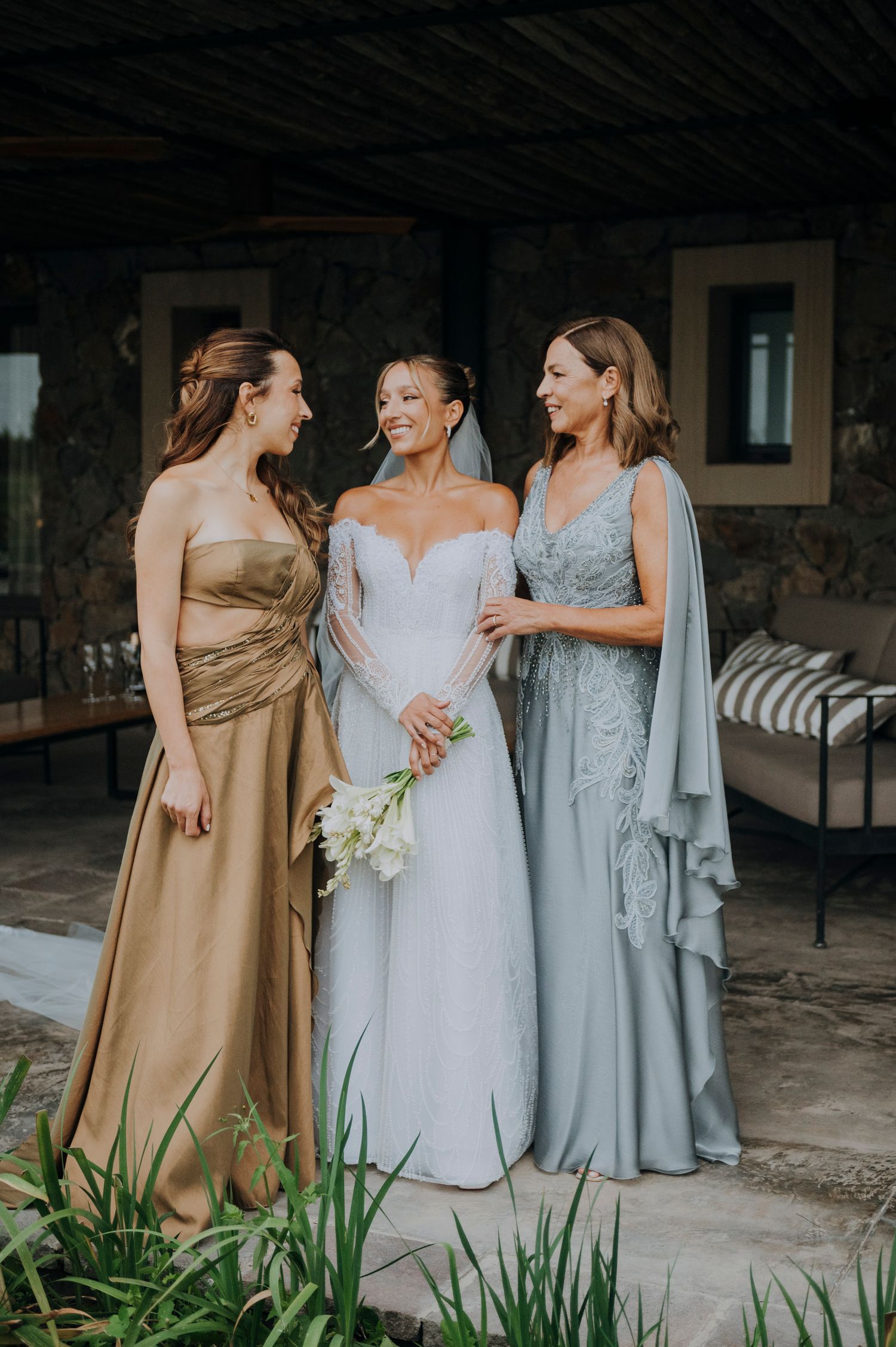 Bride in white gown poses with bridesmaid in gold and woman in blue lace dress outdoors.