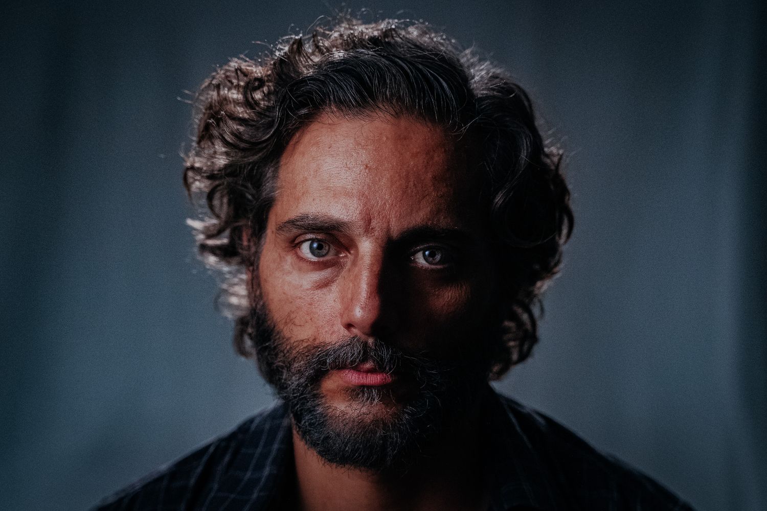 JOAQUIN FURRIEL Close-up portrait of a bearded man with curly hair against a dark background.