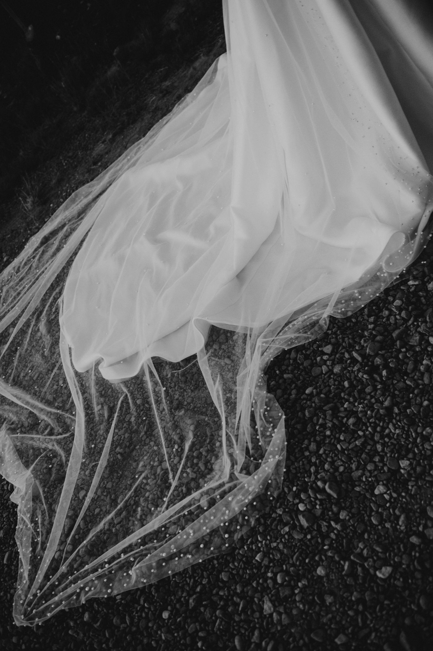 Black and white photo of a flowing wedding dress train and veil spread across a gravel surface.