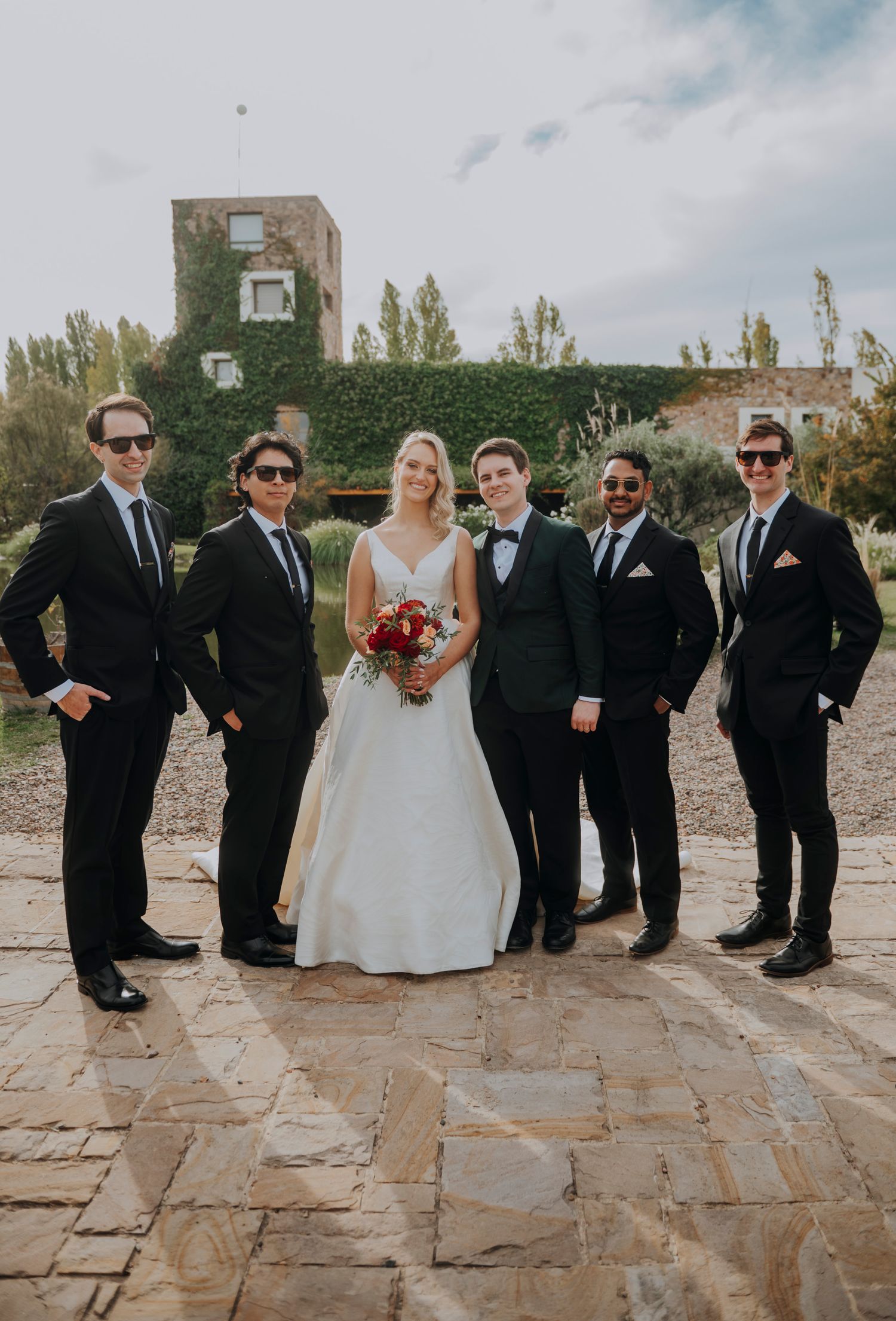 Bride in white gown poses with five groomsmen in black suits outdoors at rustic ivy-covered venue.