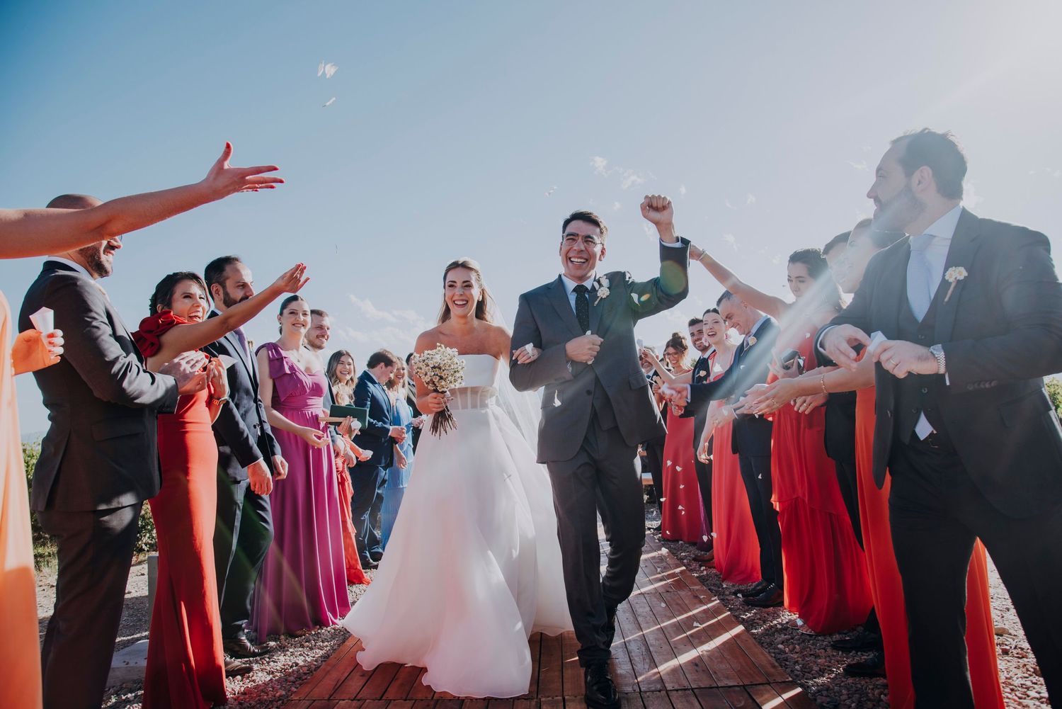 Joyful bride and groom walk through cheering wedding guests in colorful formal attire under a bright blue sky.