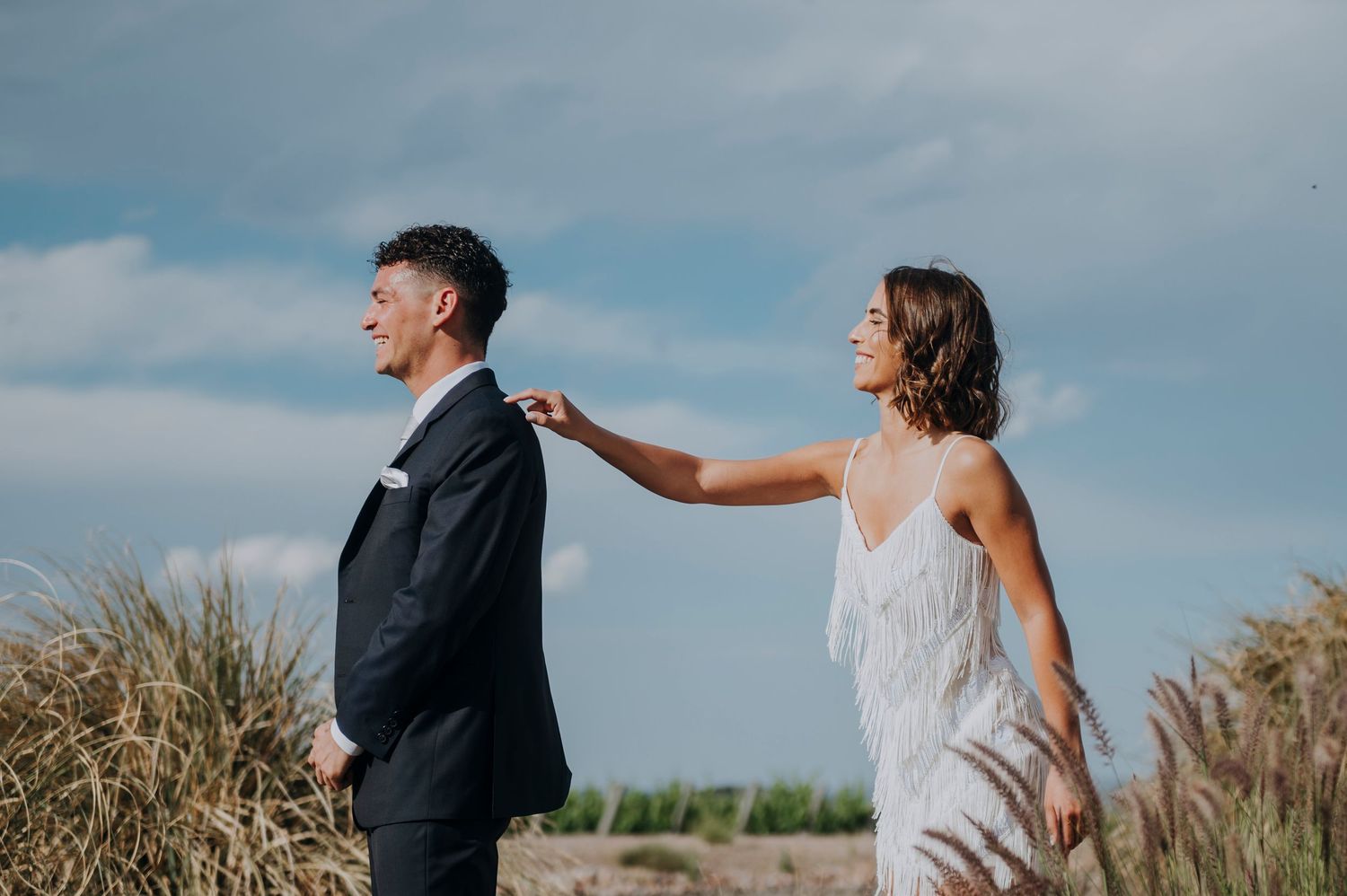 Bride in silver fringe dress taps groom on shoulder during outdoor wedding portrait in a field.