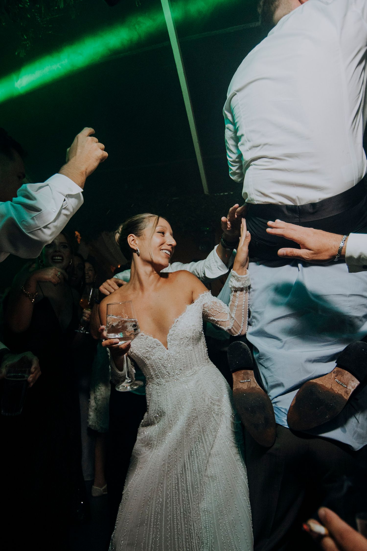 Bride in off-shoulder beaded gown smiles while dancing with guests under green lights at reception.