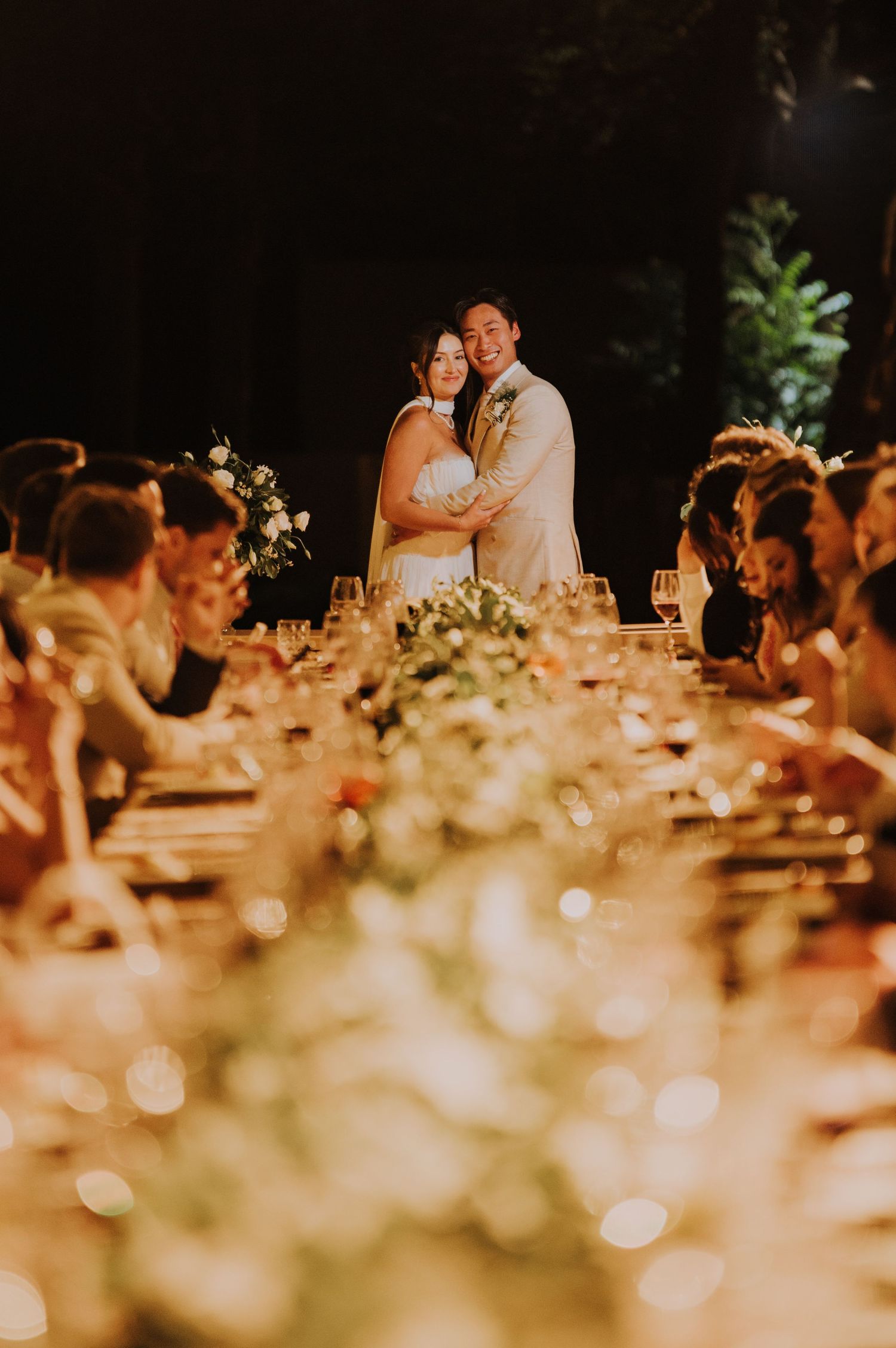 Bride and groom embracing at candlelit wedding reception with floral centerpieces.