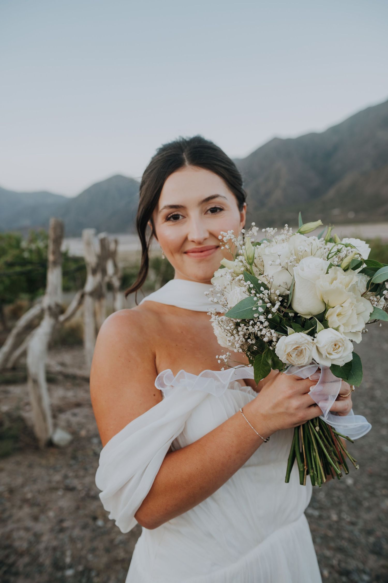 Smiling bride holds white rose bouquet in off-shoulder gown with mountain backdrop at golden hour.