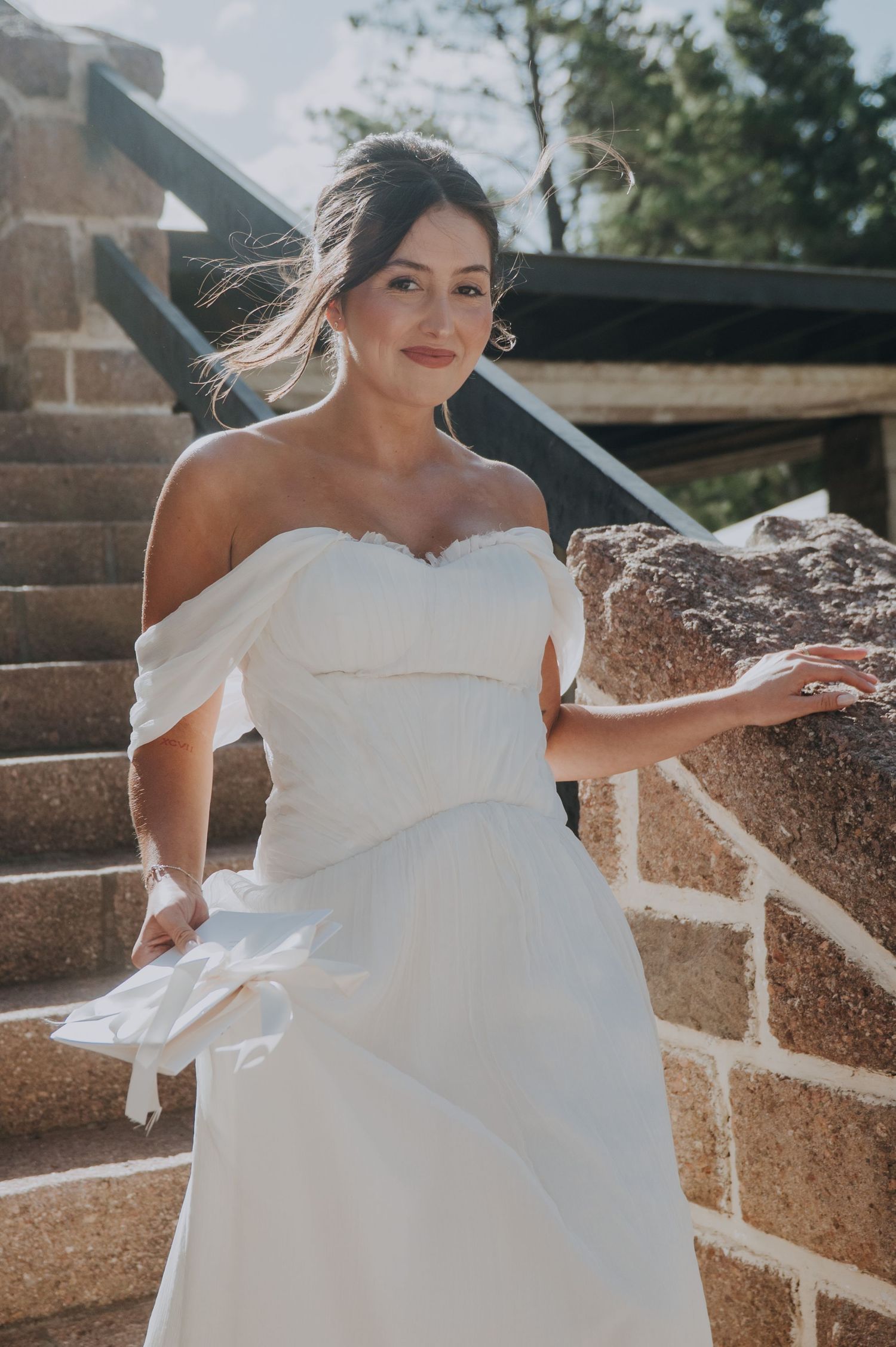 Bride in off-shoulder white sweetheart gown posing on stone steps outdoors.
