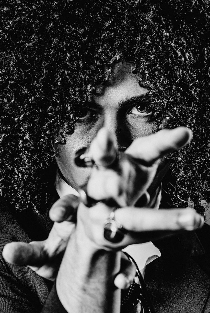 Black and white portrait of a person with curly hair reaching toward camera with ringed fingers.