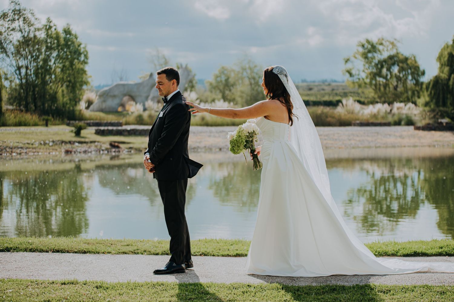 Bride touches groom's shoulder during first look moment beside a serene reflective pond.