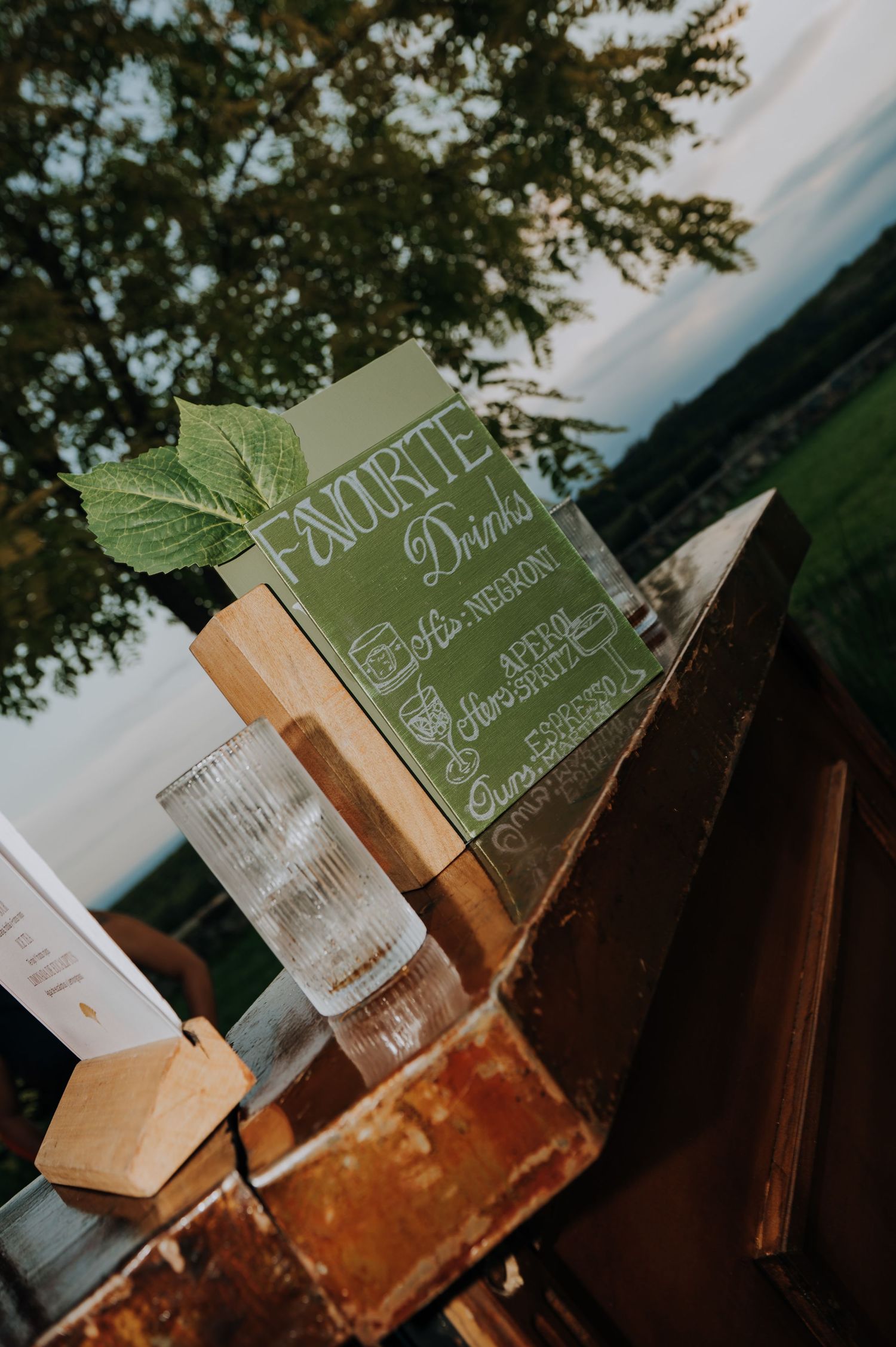 Rustic wedding bar sign listing favorite drinks displayed on wooden surface at outdoor evening reception.