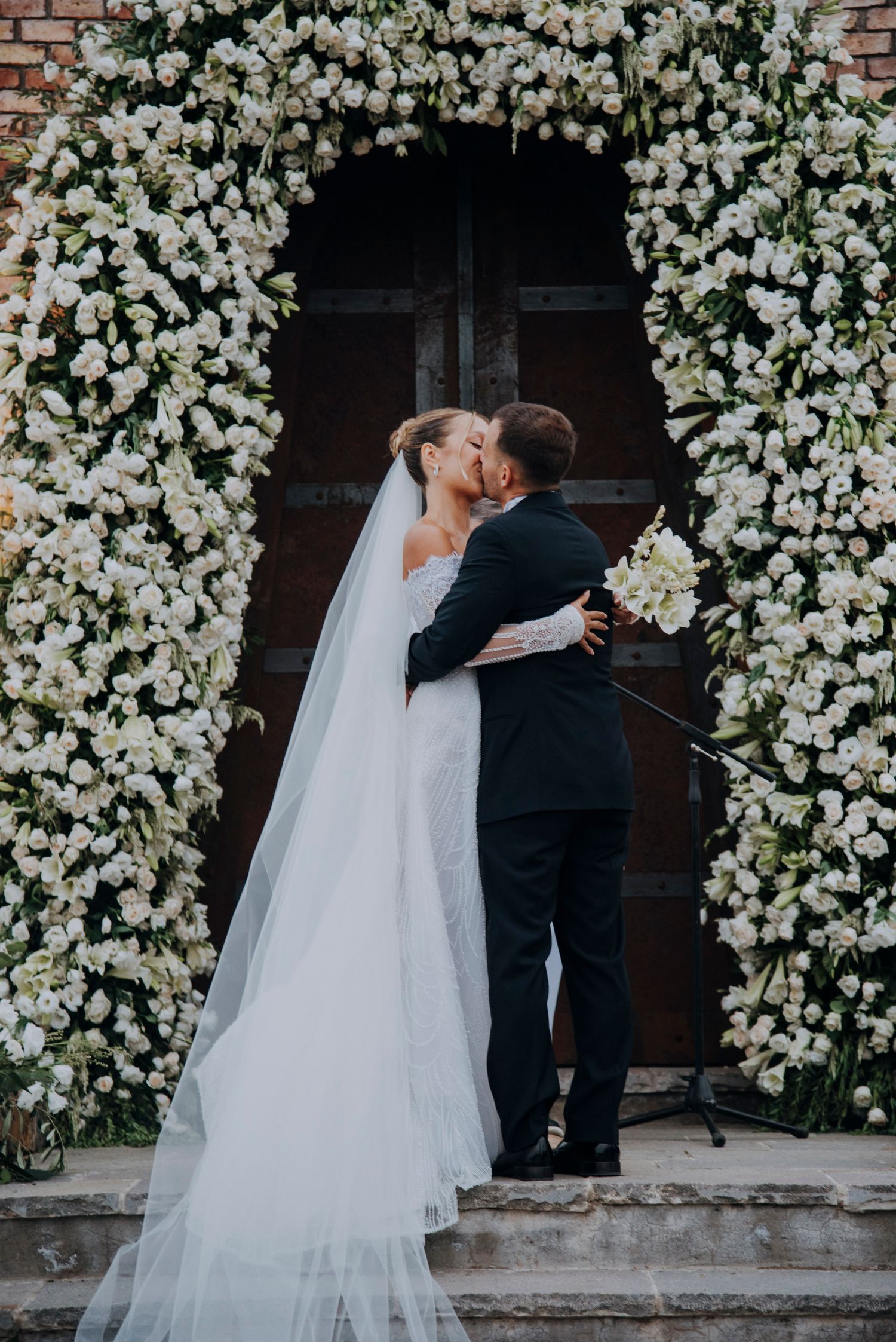 Bride and groom share first kiss under stunning white floral arch at church wedding ceremony.