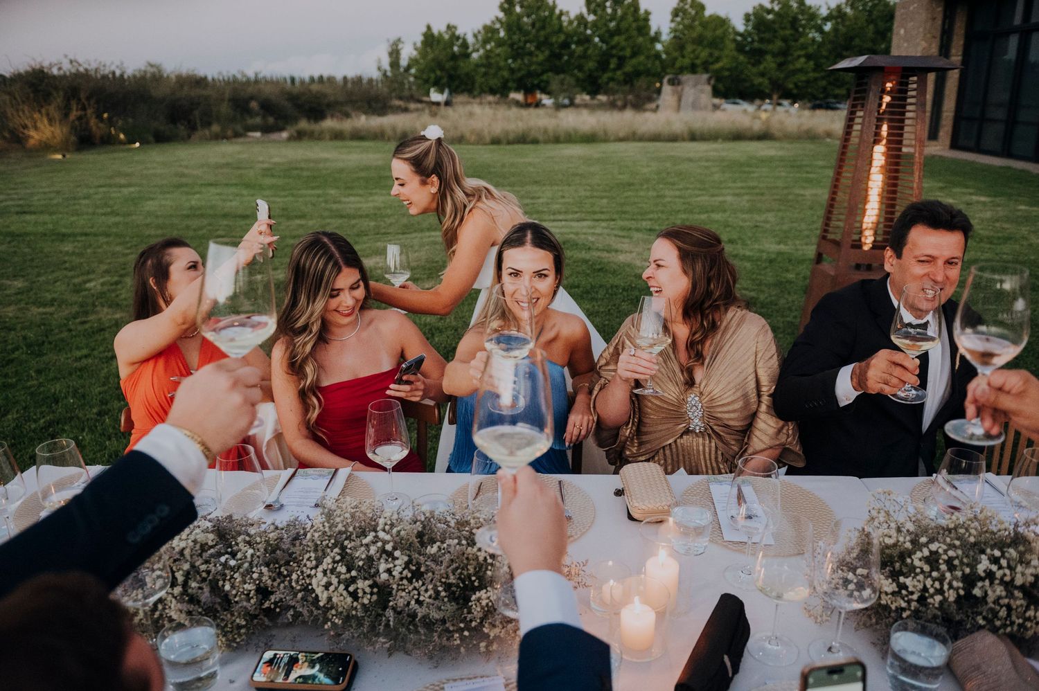 Bridesmaids and guests raise glasses in a toast at elegantly decorated outdoor wedding dinner table.