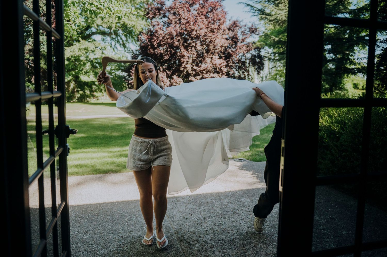 Woman carries a flowing white wedding veil through open French doors in a sunny outdoor setting.
