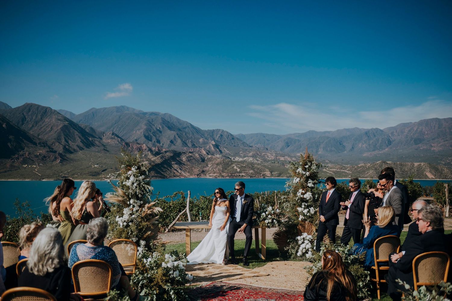 Outdoor wedding ceremony with mountain and lake backdrop, bride and groom at altar, guests seated in rustic chairs.