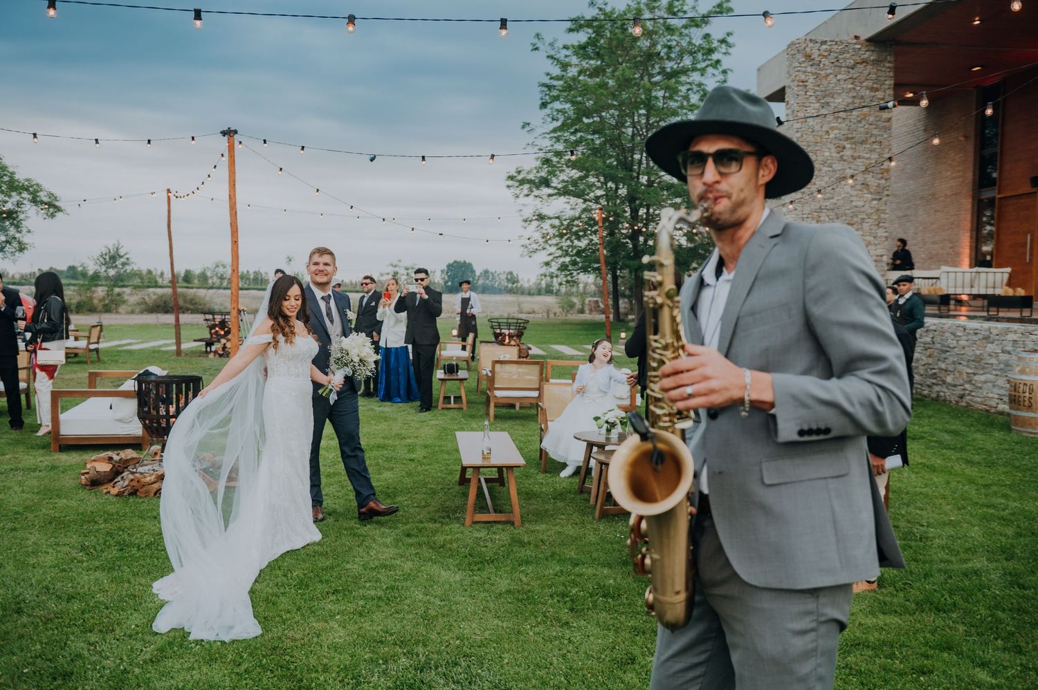 Saxophonist in gray suit and hat performs at outdoor wedding ceremony while bride and groom walk on green lawn.