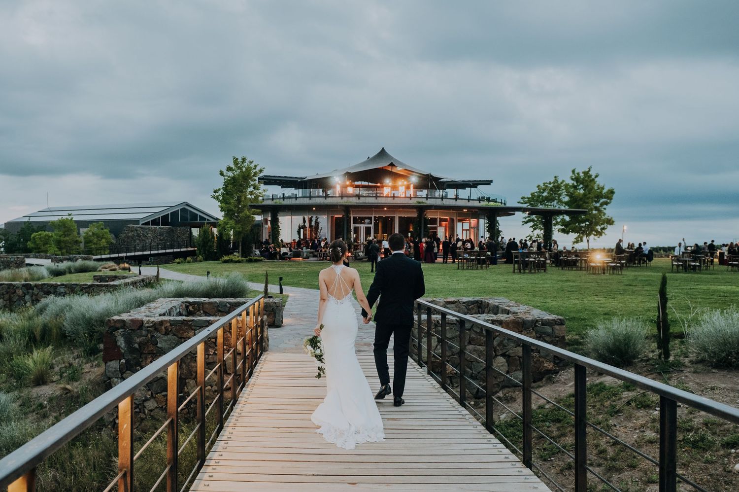Bride and groom walk hand-in-hand on a wooden bridge toward a lit wedding venue under cloudy skies.