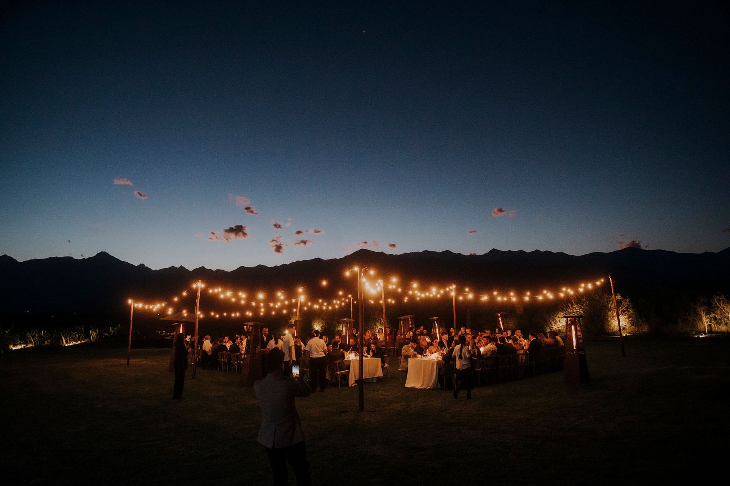 Magical outdoor wedding reception under string lights with mountain backdrop at twilight.