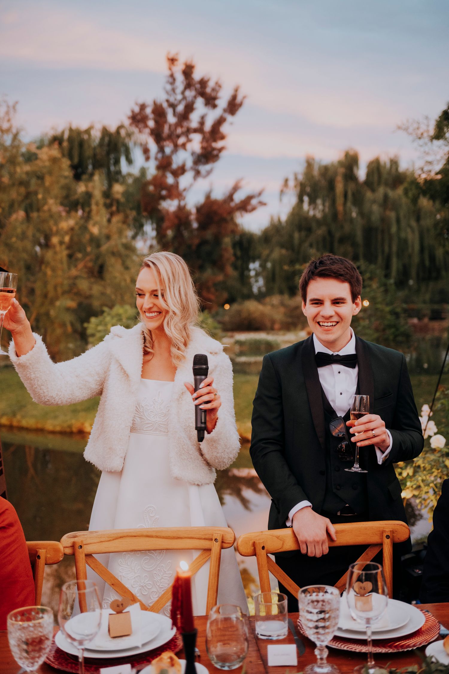 Smiling bride in white fur stole and groom in tuxedo raising champagne glasses during outdoor reception toast.