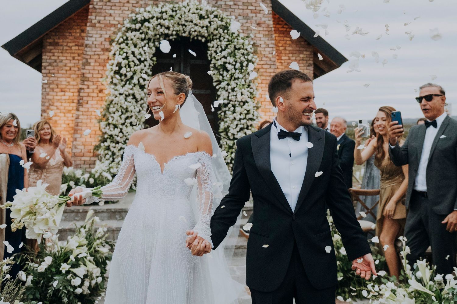 Joyful newlyweds walk hand-in-hand through floral arch at outdoor wedding ceremony with guests celebrating.