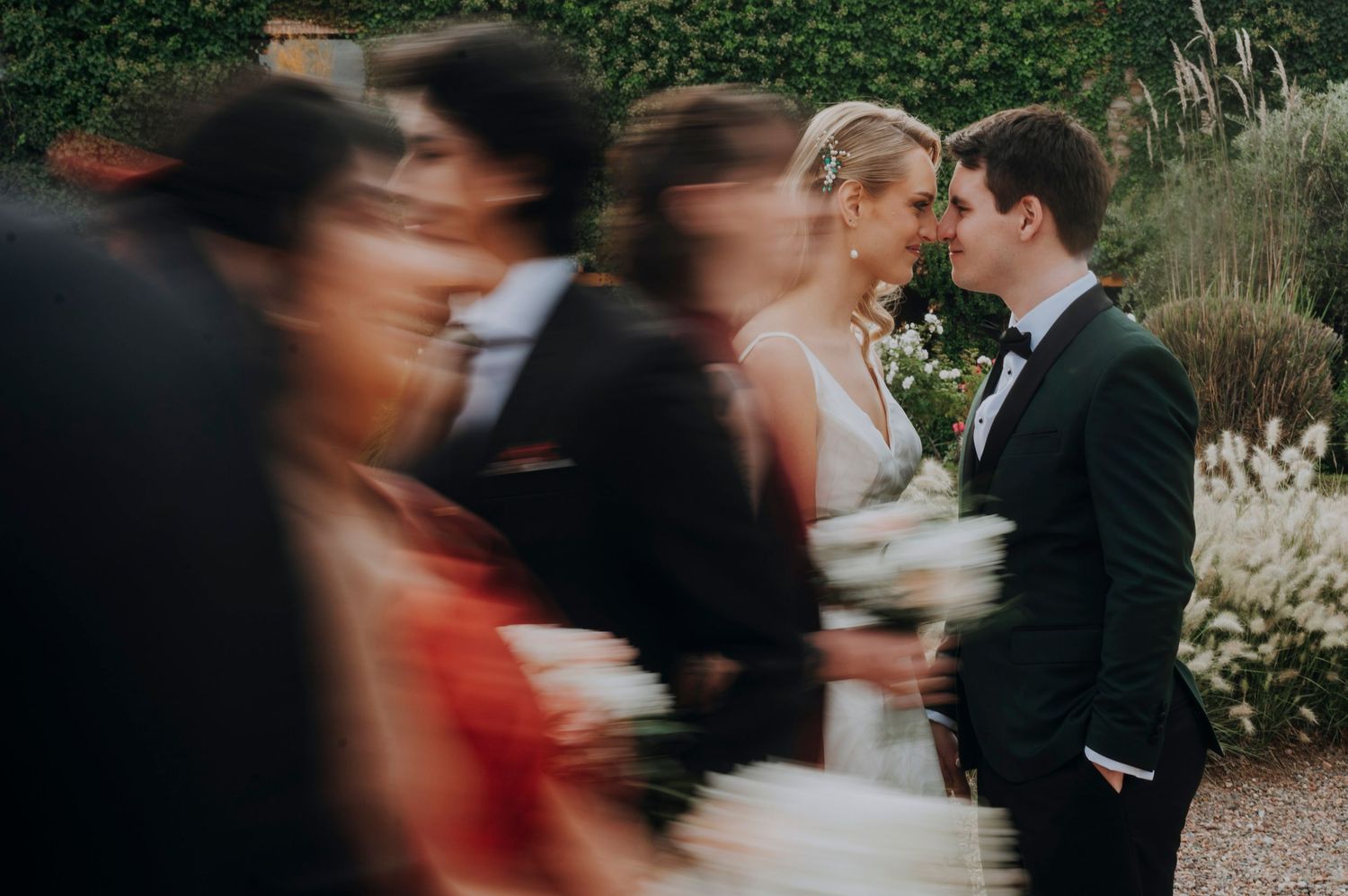 A bride and groom share an intimate moment while wedding guests blur past them in motion at an outdoor garden ceremony.