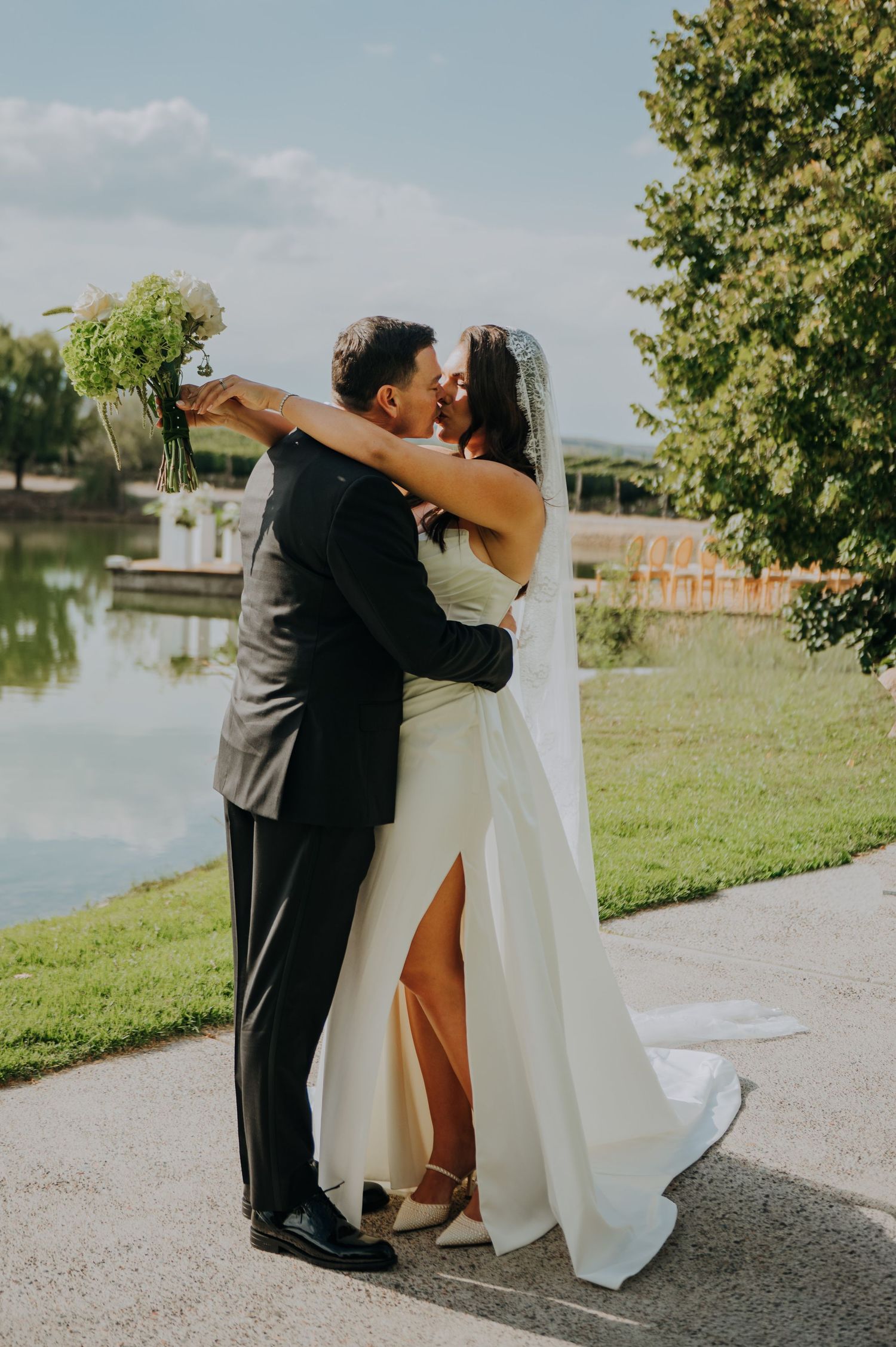 Bride and groom share a romantic kiss outdoors by a serene lake on their wedding day.