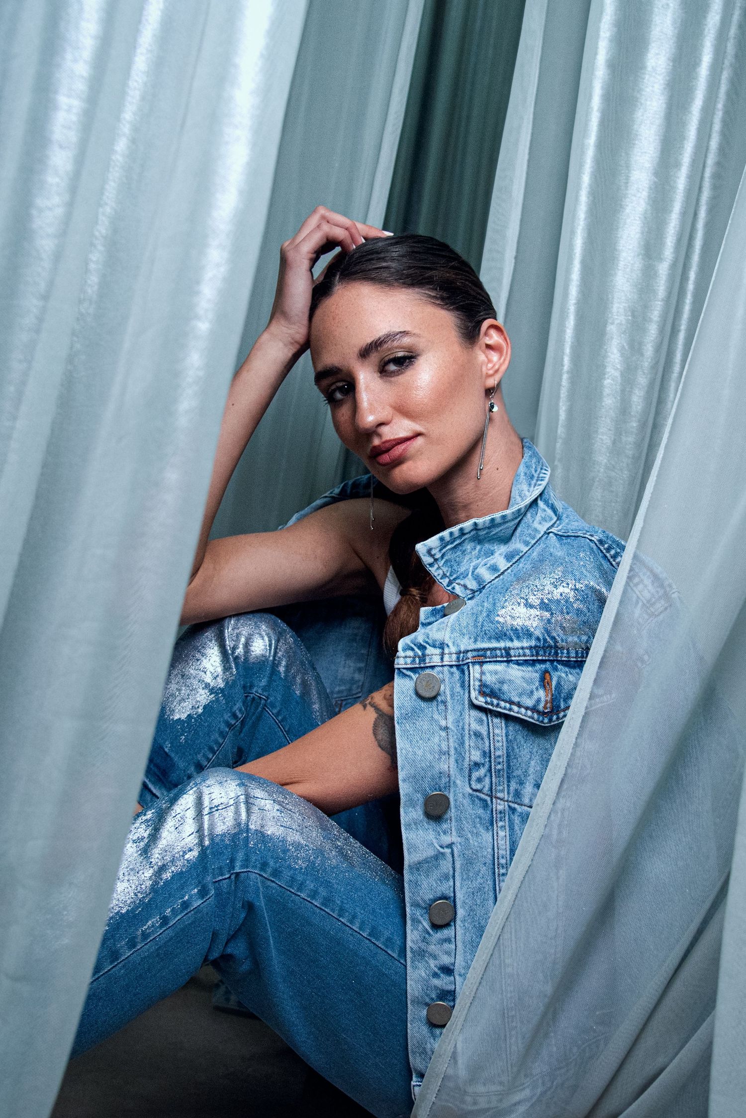 Woman in denim jacket and jeans sitting between curtains with a tattoo.