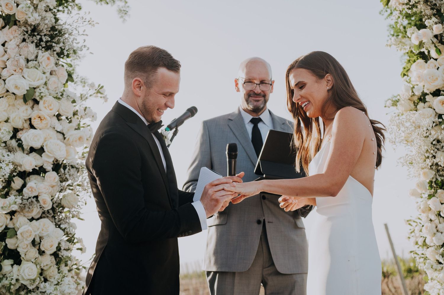 Groom places ring on bride's finger under floral arch as officiant watches during outdoor wedding ceremony.