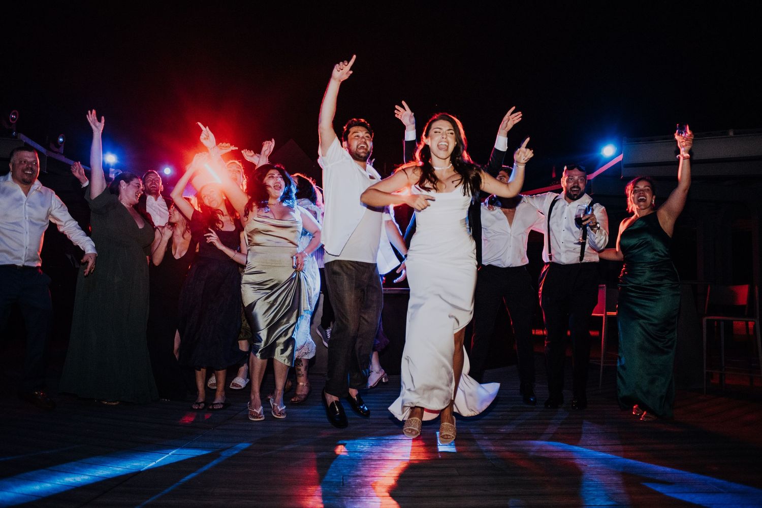 Bride and groom jump joyfully on the dance floor surrounded by celebrating wedding guests.